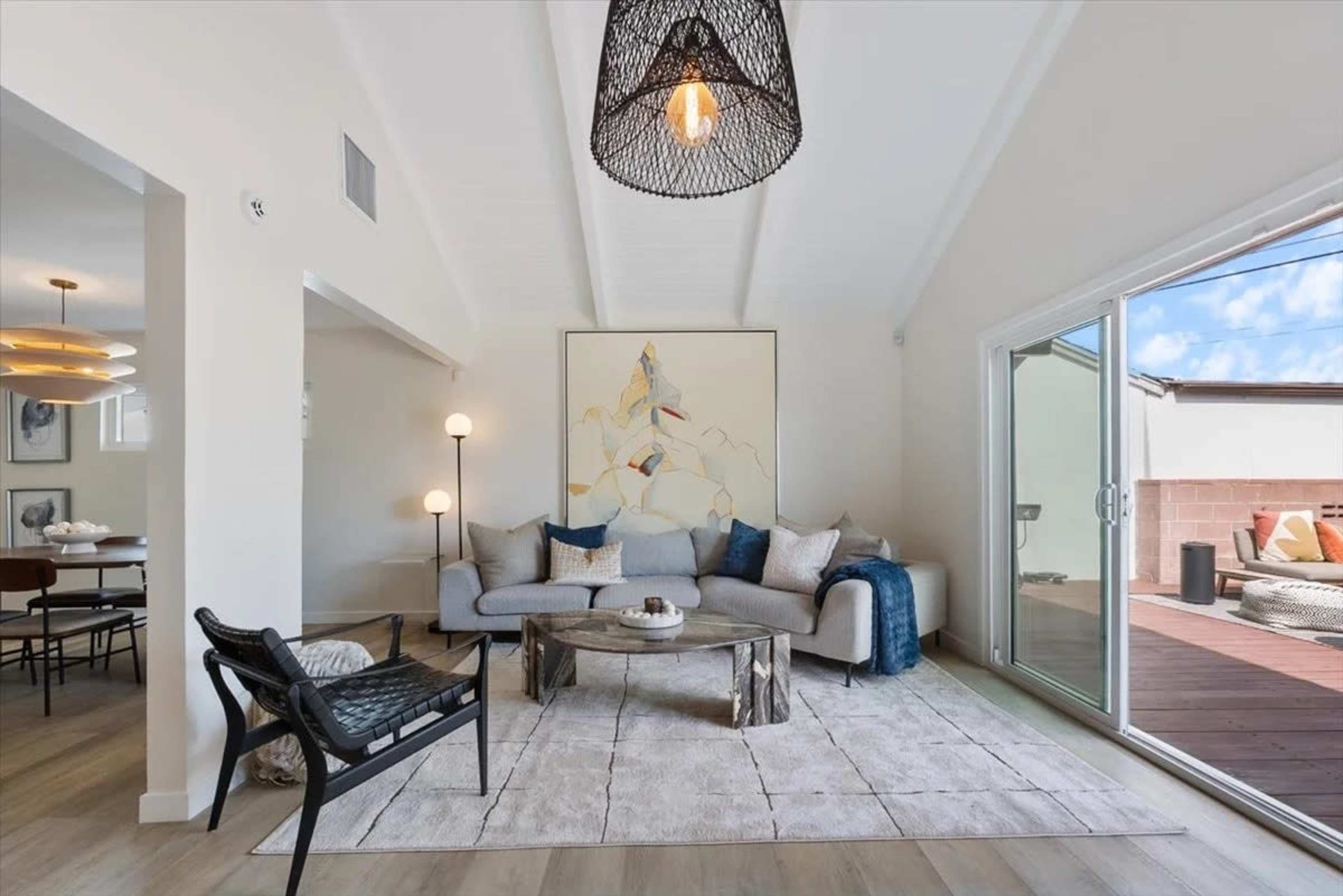 A bright and airy living room features a gray sectional sofa, a circular wooden coffee table, and a large artwork above the sofa, with sliding glass doors leading to an outdoor deck.