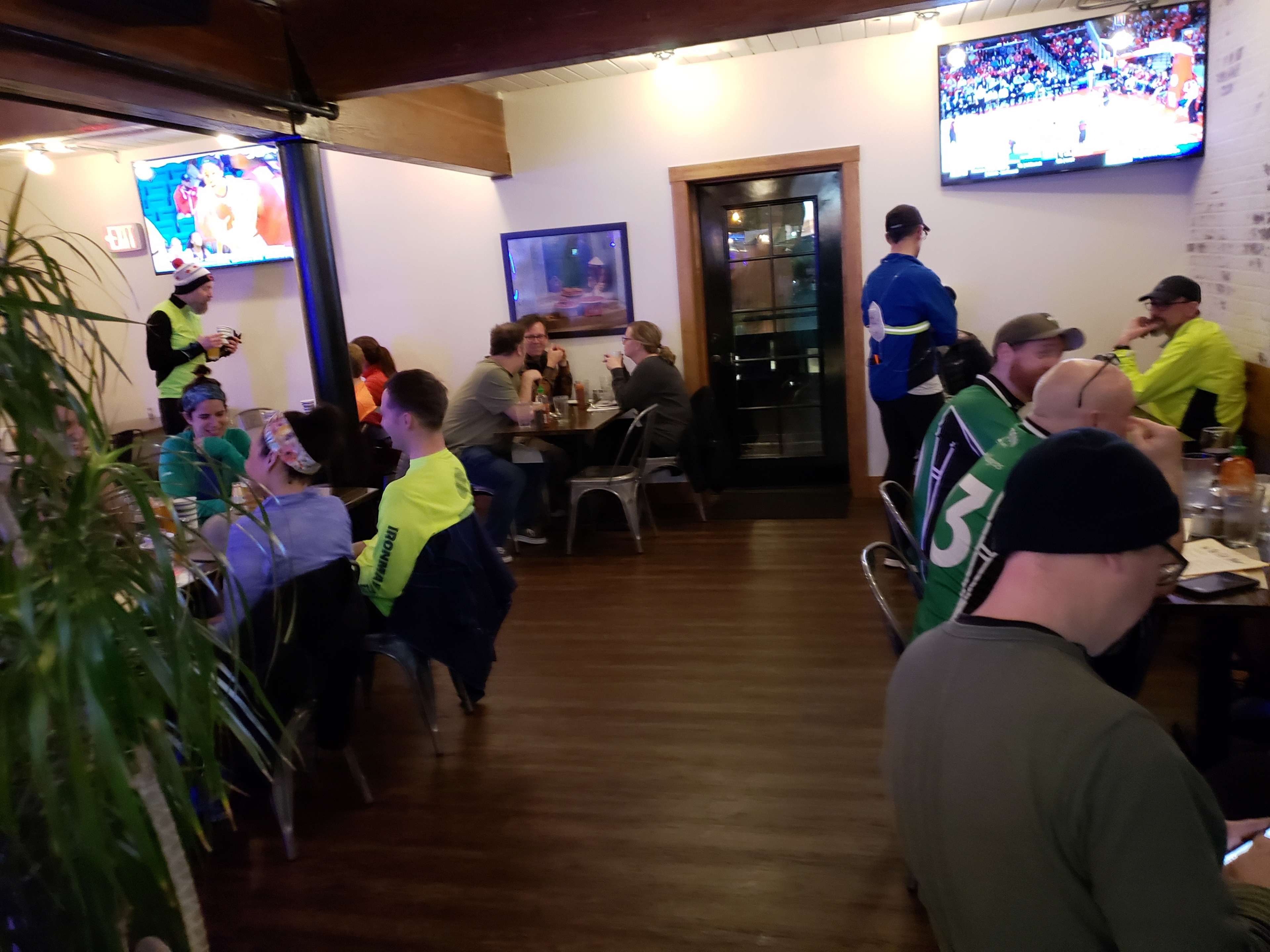 A restaurant interior with patrons sitting at tables, a staff member taking an order, and multiple televisions displaying a sports event.