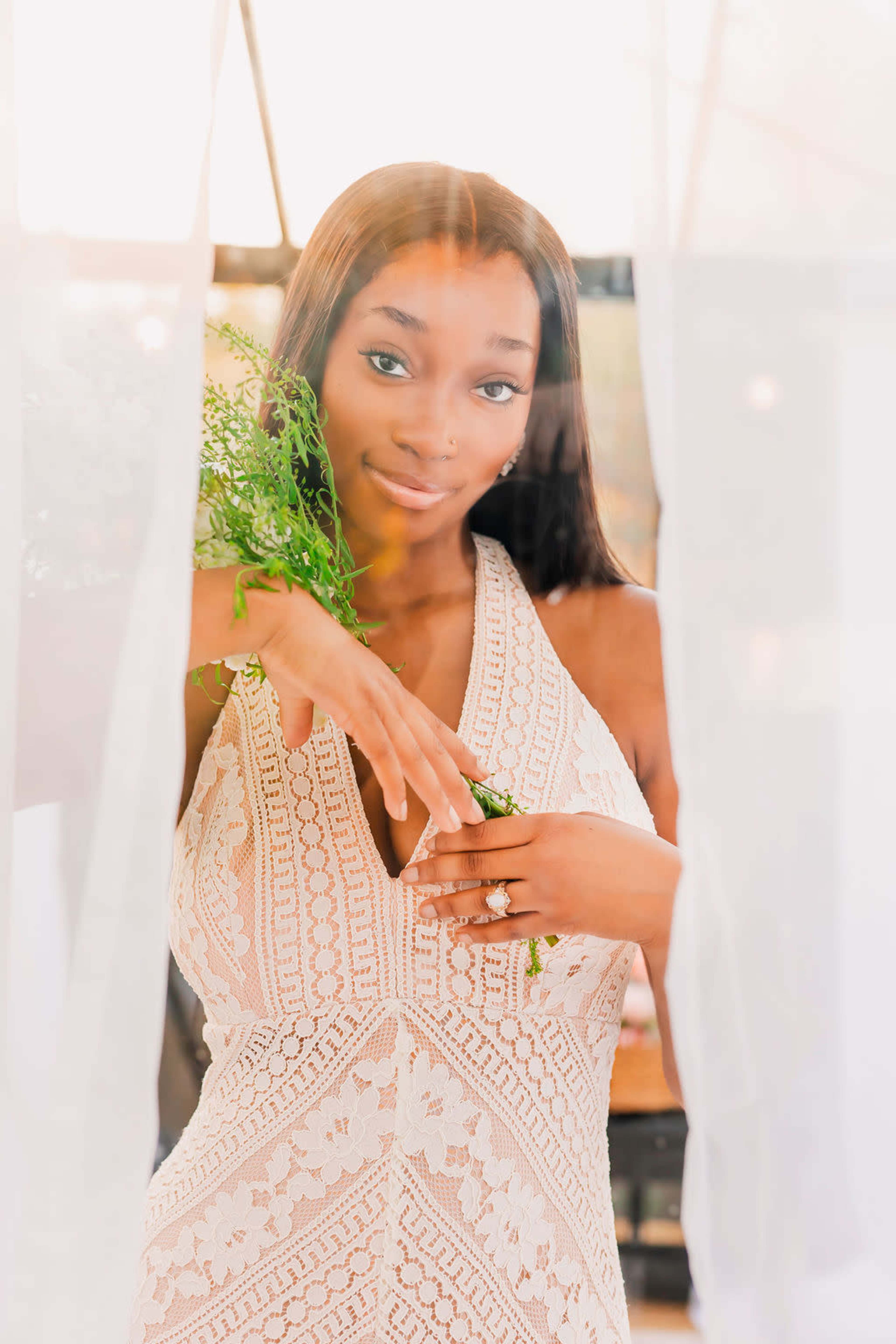 A woman in a lace dress stands holding greenery, partially framed by sheer curtains.