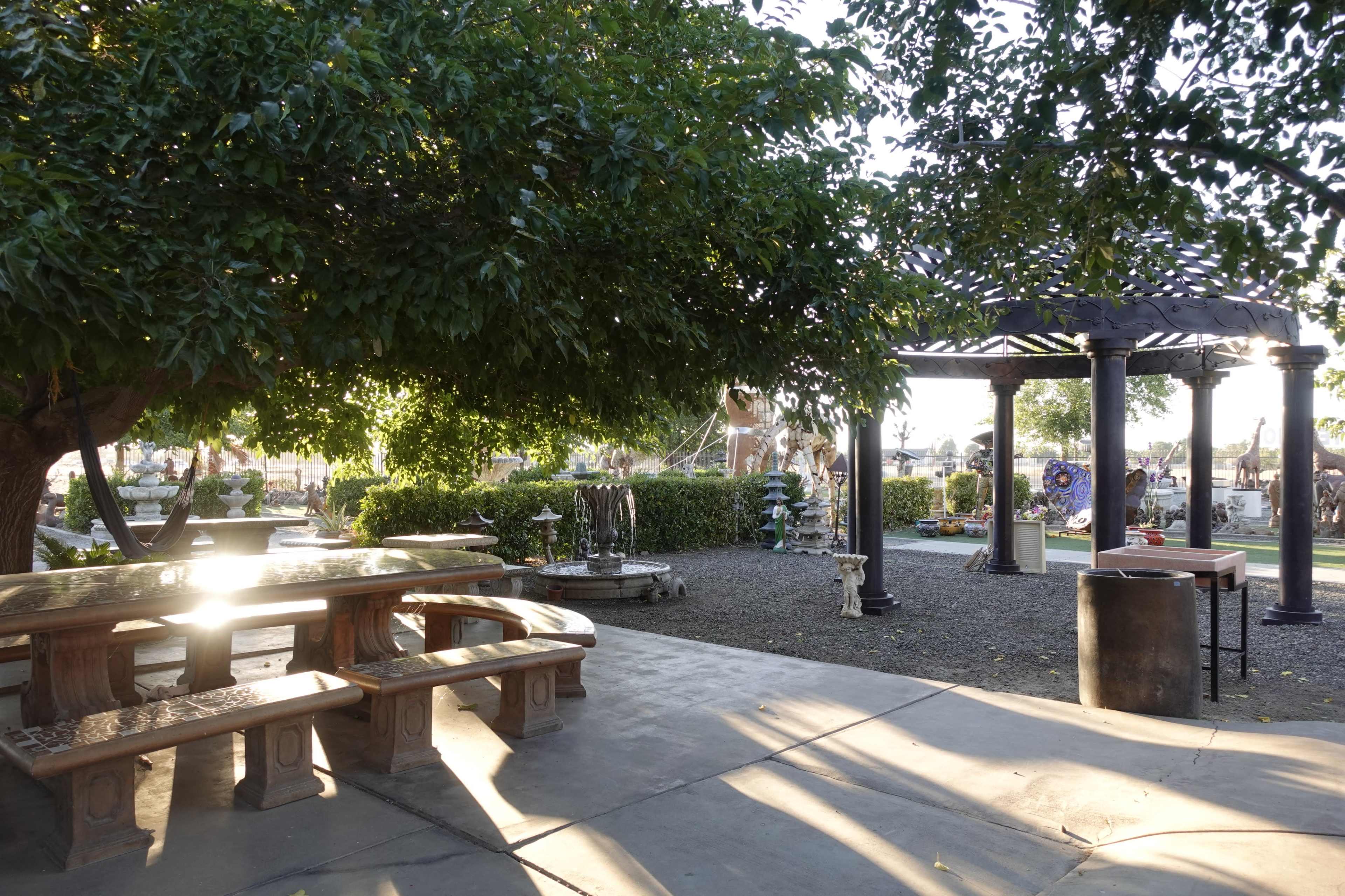 The scene depicts an outdoor area with stone tables and benches under a large tree, surrounded by sculptures and gardens near a pergola.