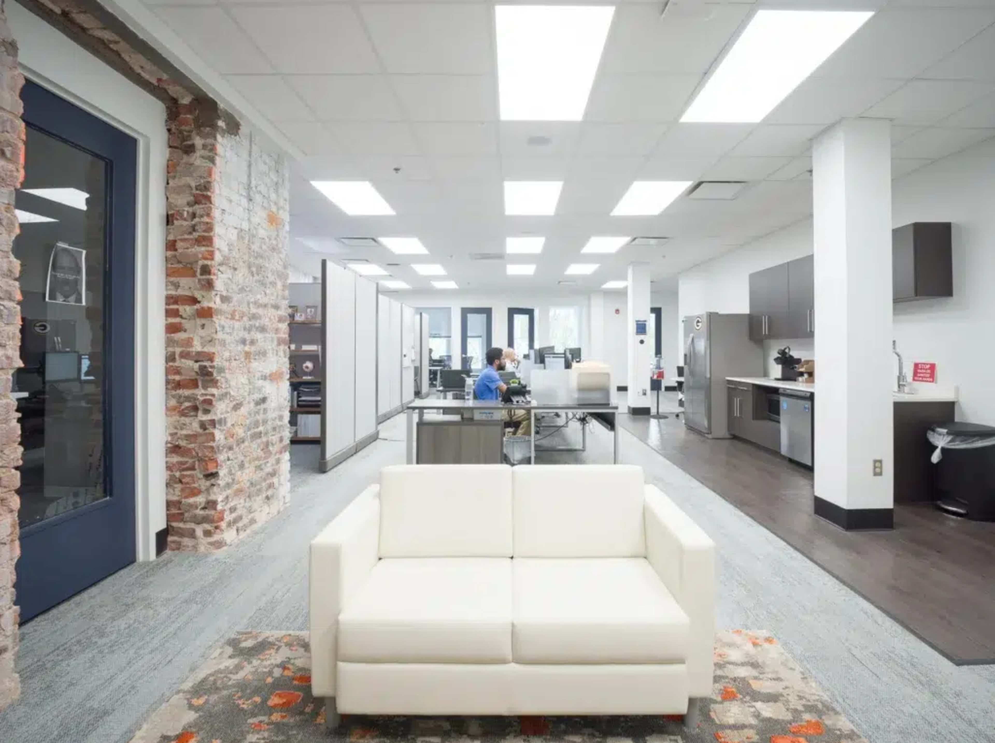 The image shows a modern office space featuring a white sofa in the foreground, with workstations and kitchen areas visible in the background.