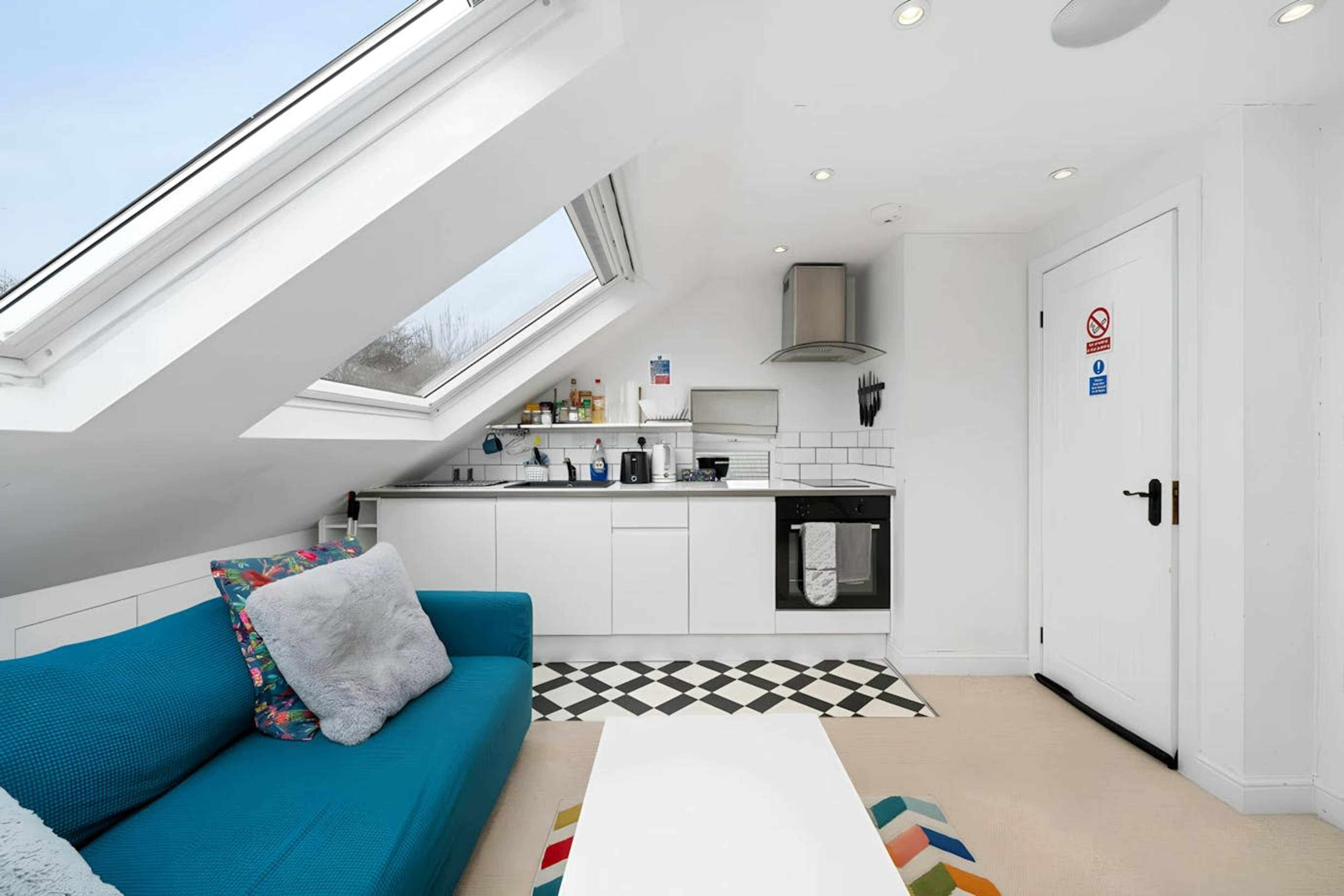 The image shows a small, modern kitchen area with a sloped ceiling, featuring a window and a sofa, along with a black and white checkered floor.