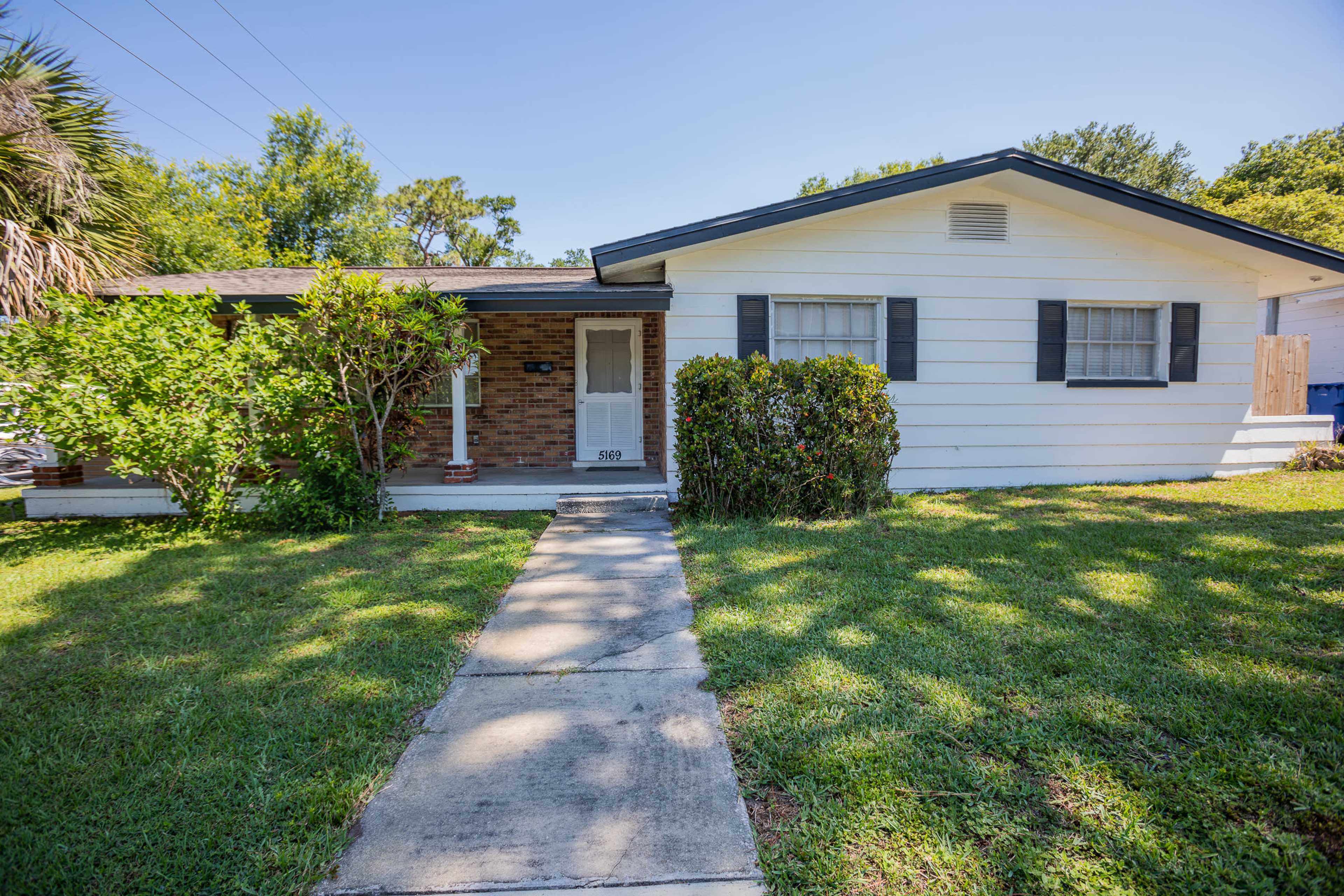 The image shows a single-story house with a brick and white exterior, a front walkway, and a well-maintained lawn.