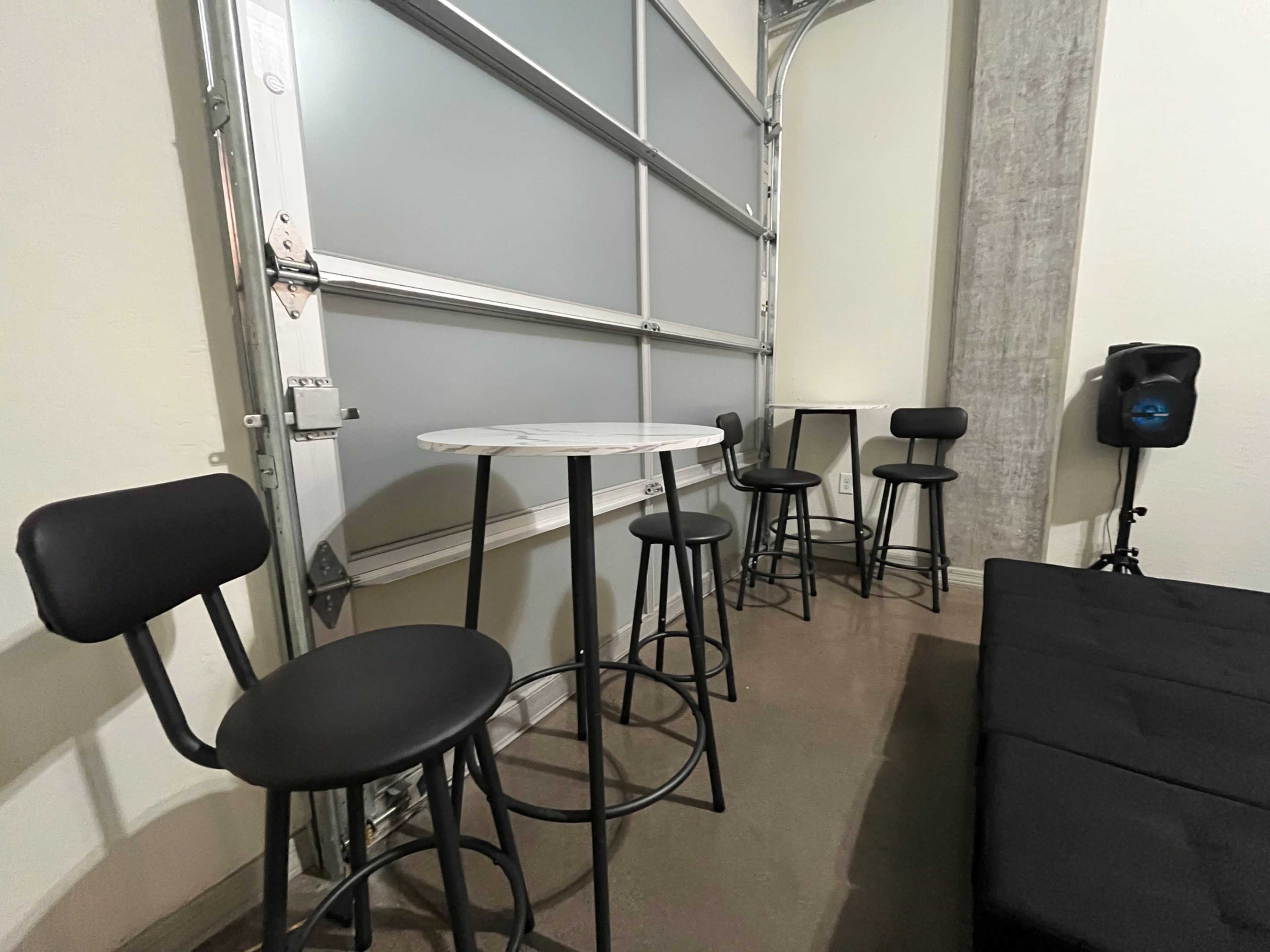 The image shows a small seating area with a high round table and four black bar stools near a garage door.