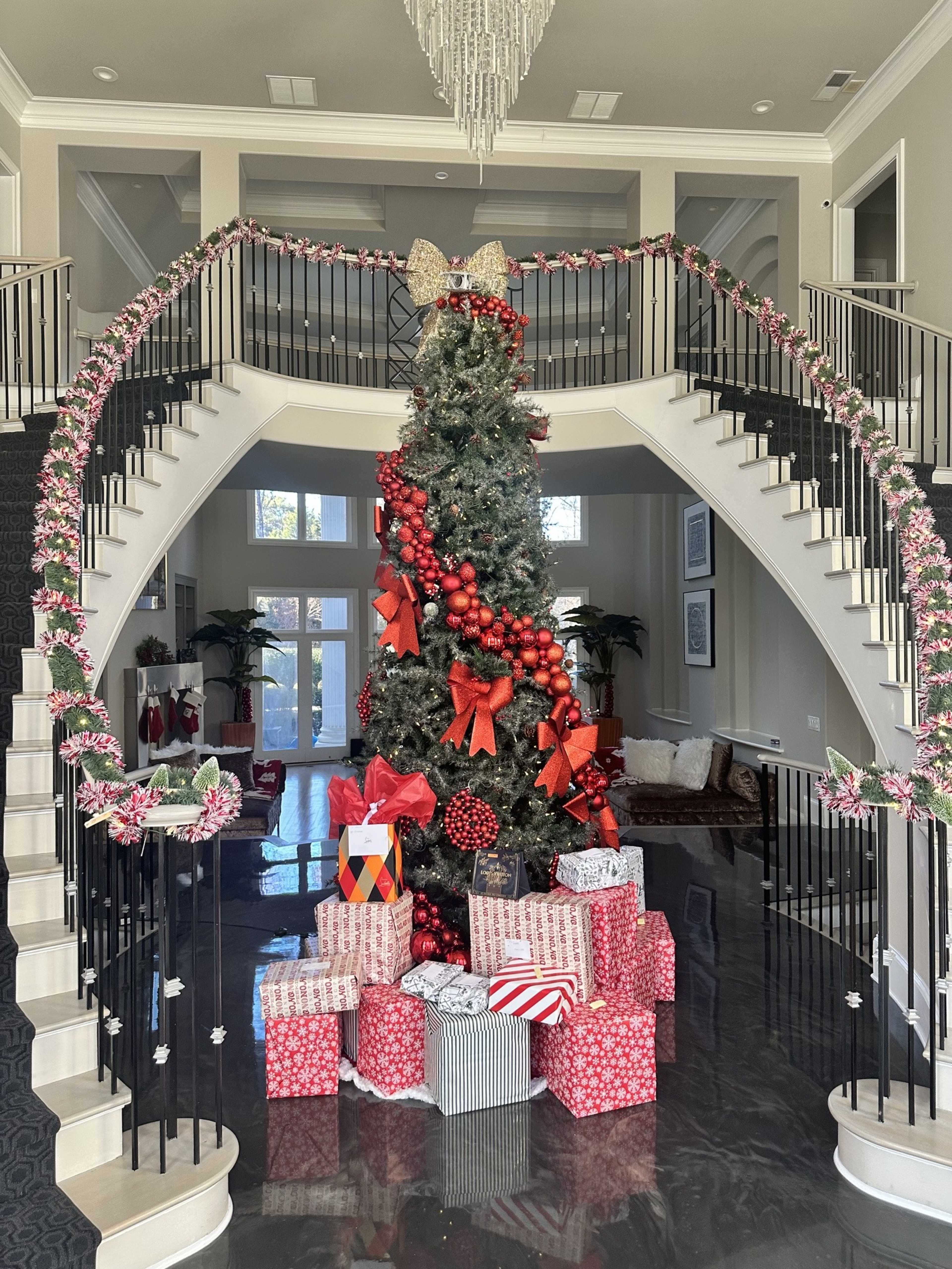 A beautifully decorated Christmas tree with red and gold ornaments stands at the base of a grand staircase, surrounded by an array of wrapped gifts.
