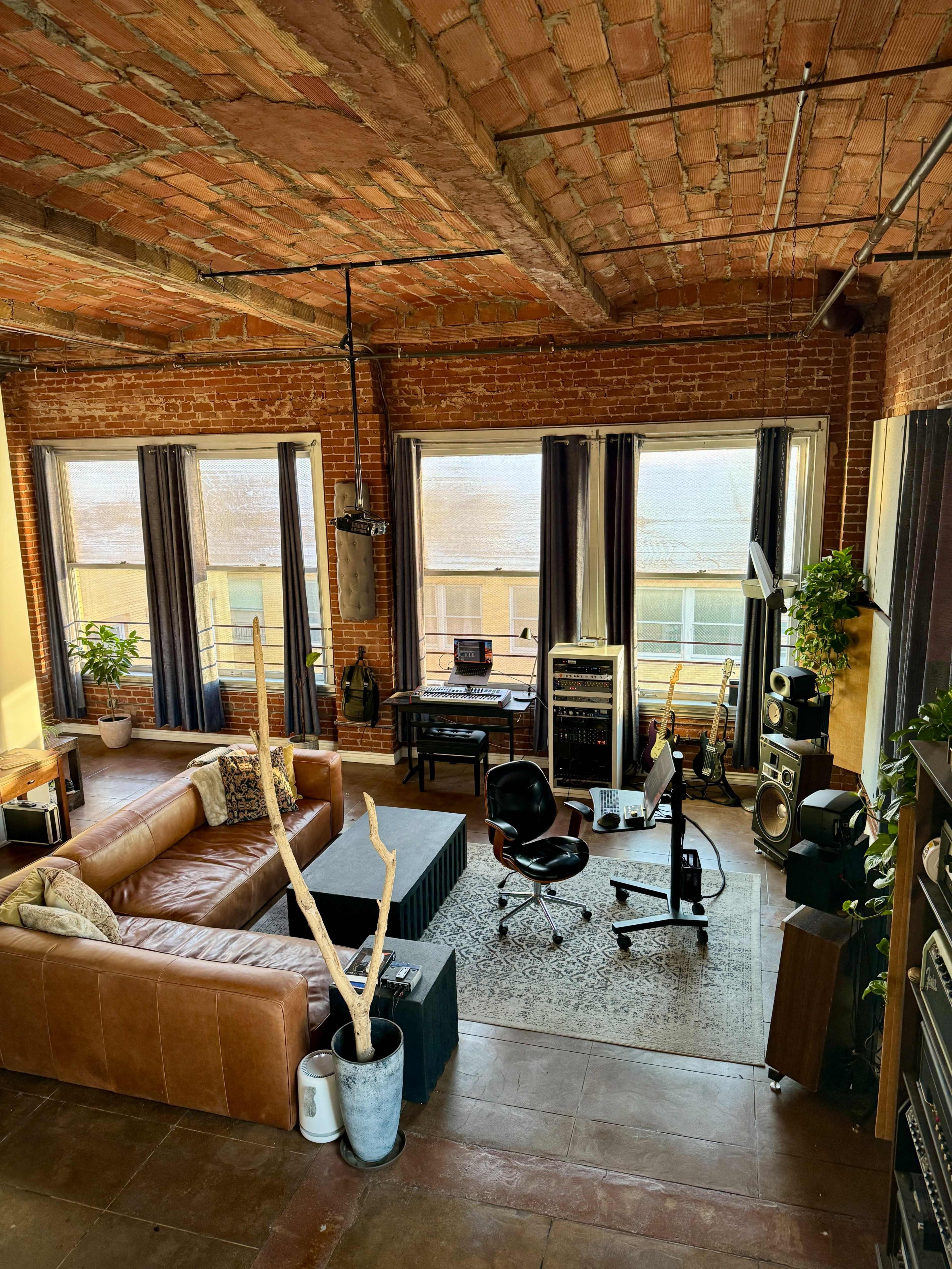 The image shows a spacious loft living room with exposed brick walls, large windows, a leather sofa, a coffee table, and a home studio setup featuring audio equipment.