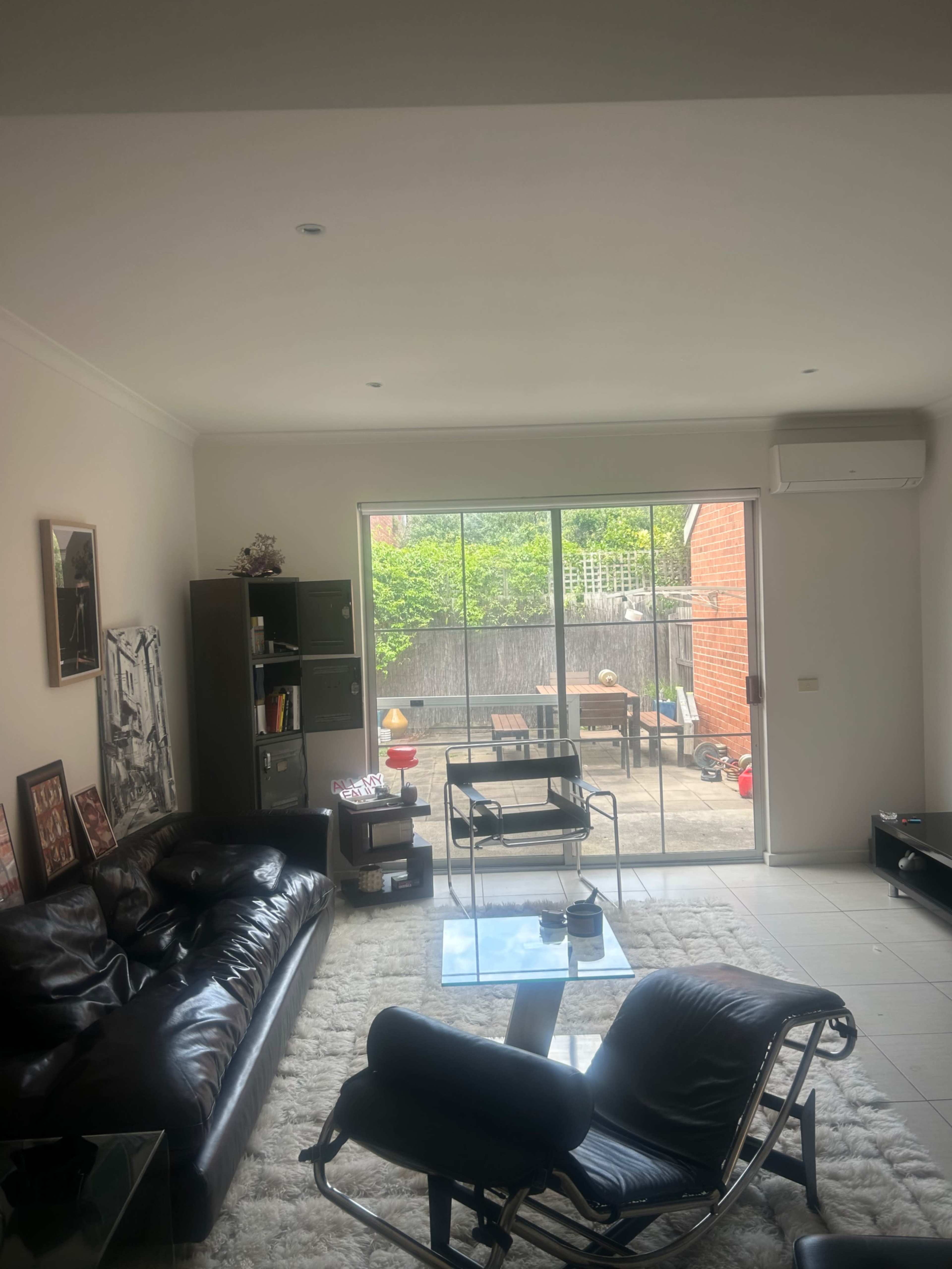 A minimalist living room features a black leather sofa, a glass coffee table, and a view of a patio with outdoor furniture through large sliding doors.