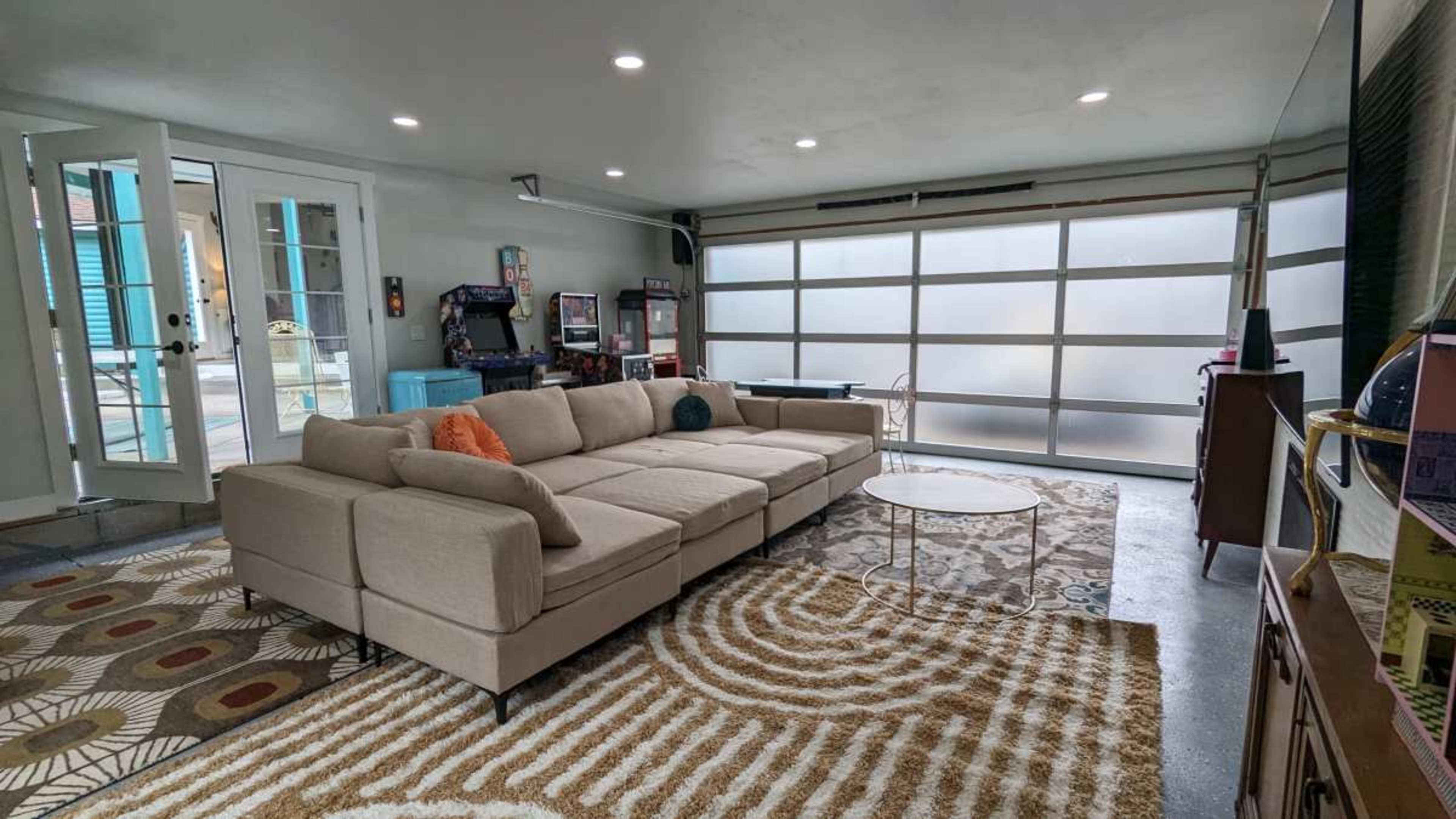 The image shows a modern living area featuring a large sectional sofa, a round coffee table, a patterned rug, and a garage door window allowing natural light.