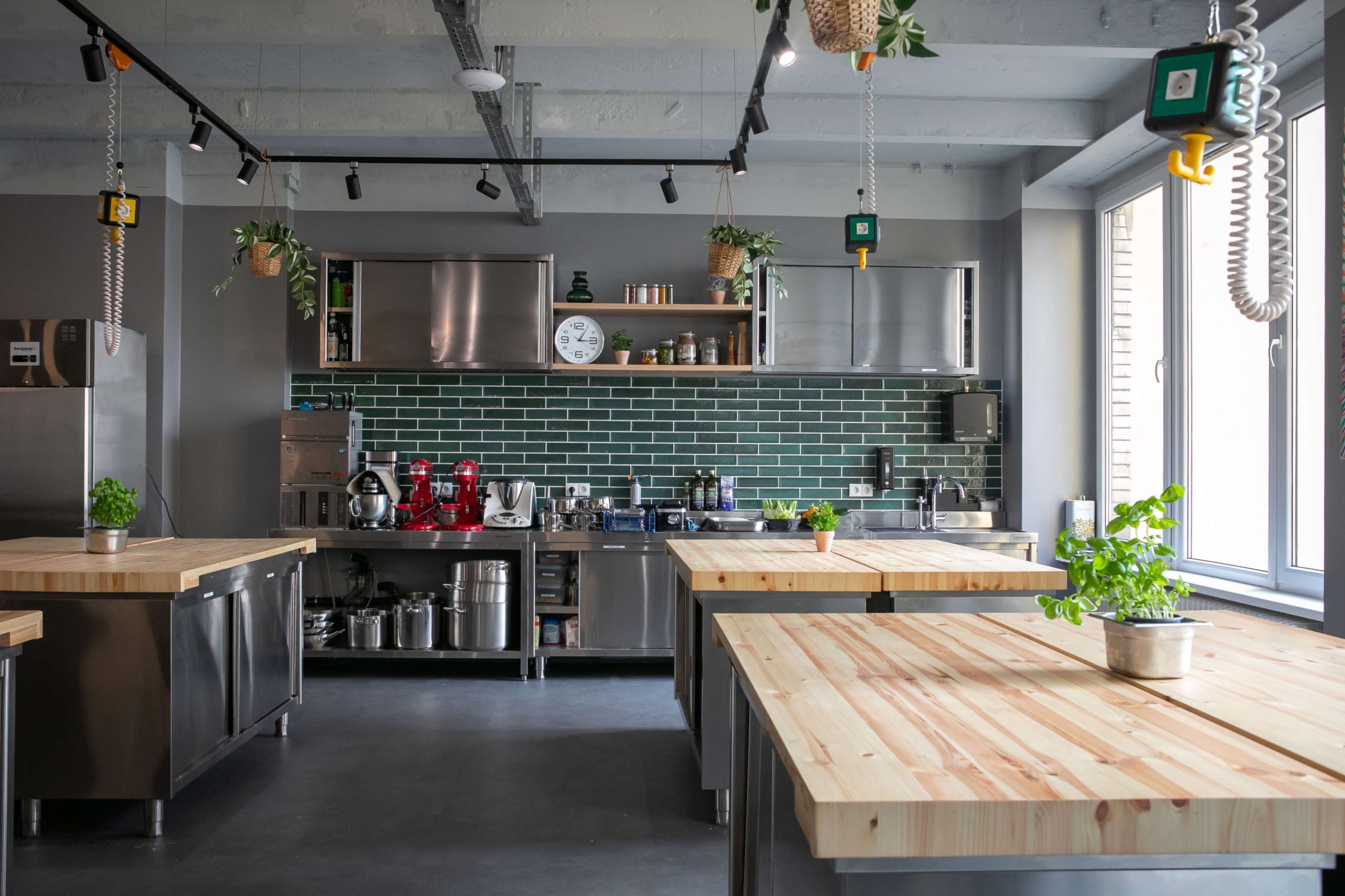 The image shows a modern kitchen with stainless steel appliances, wooden countertops, and green tile backsplash, featuring several hanging plants.