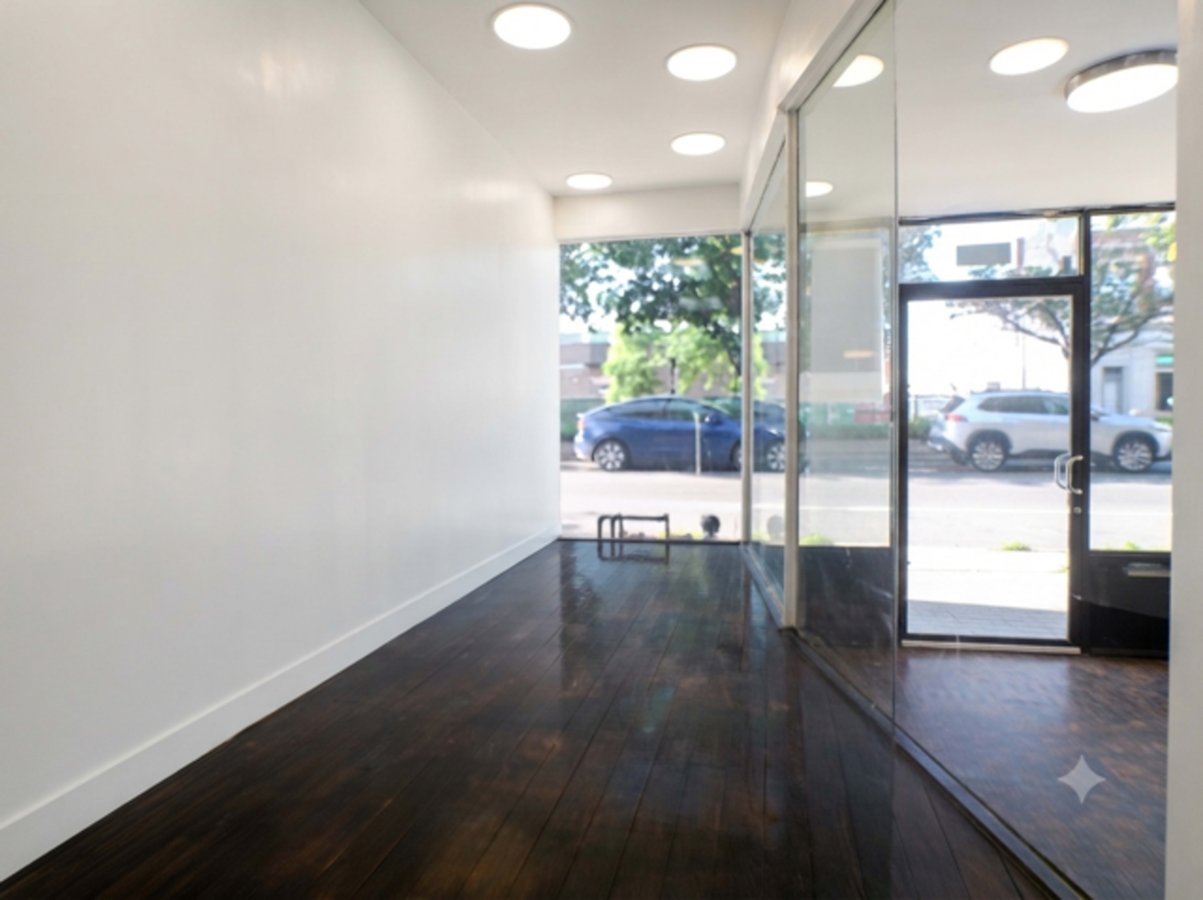 The image shows a spacious, minimalist interior featuring a wooden floor and large glass windows that allow natural light to enter.