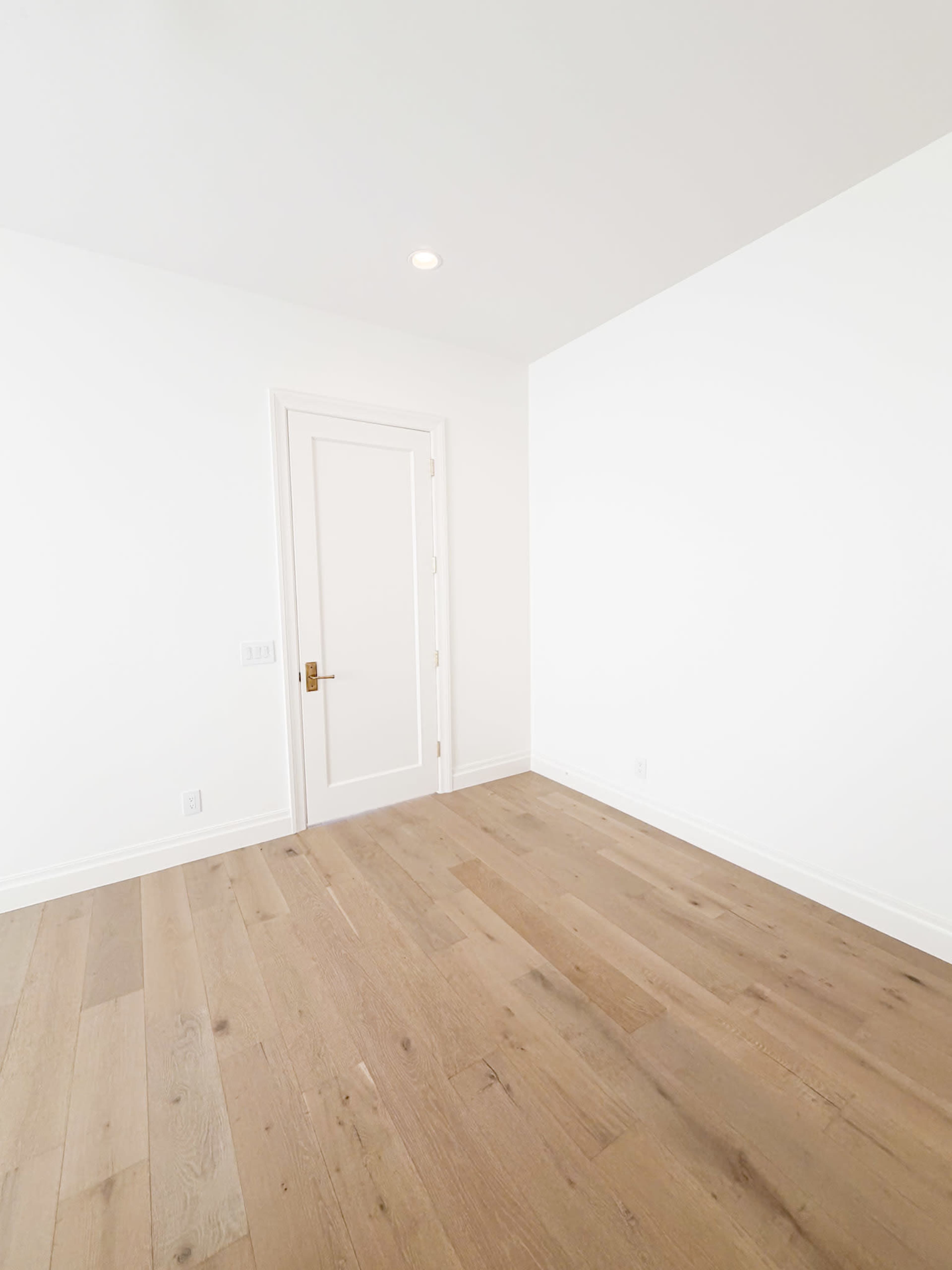 The image shows a minimally furnished room with light wooden floors and a white wall featuring a closed door.
