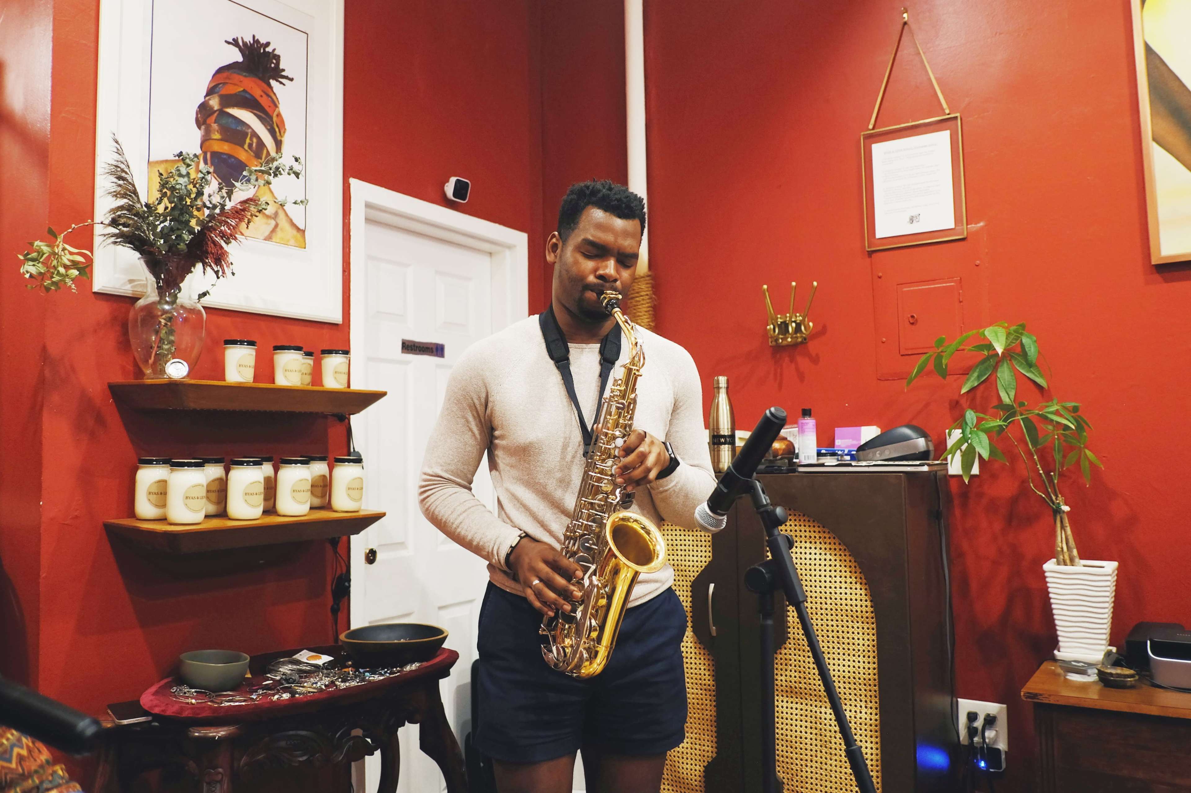 A man plays the saxophone in a room with red walls, surrounded by artwork and decorative plants.