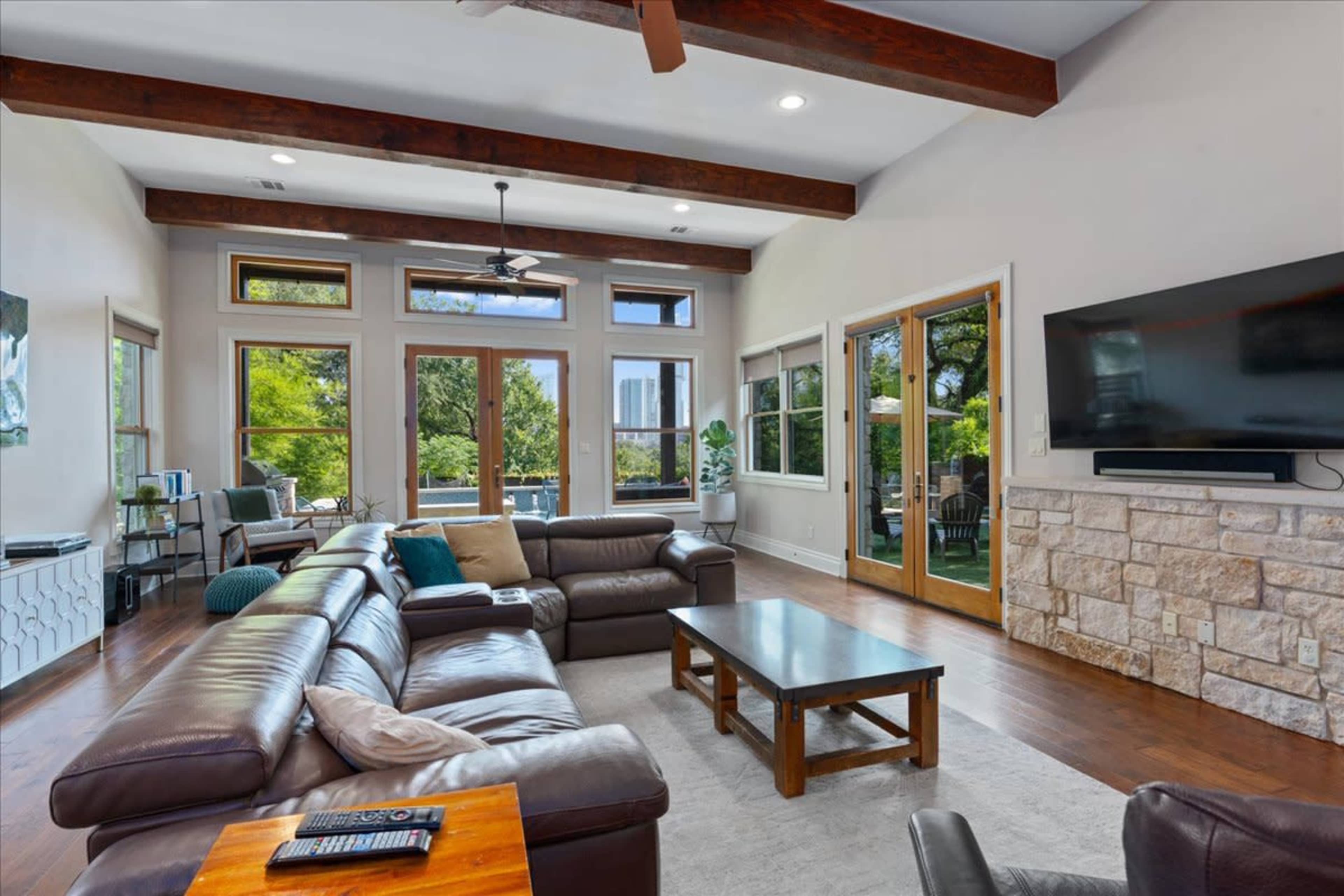 A spacious living room features a leather sectional sofa, a coffee table, large windows with views, and a mounted television on a stone wall.