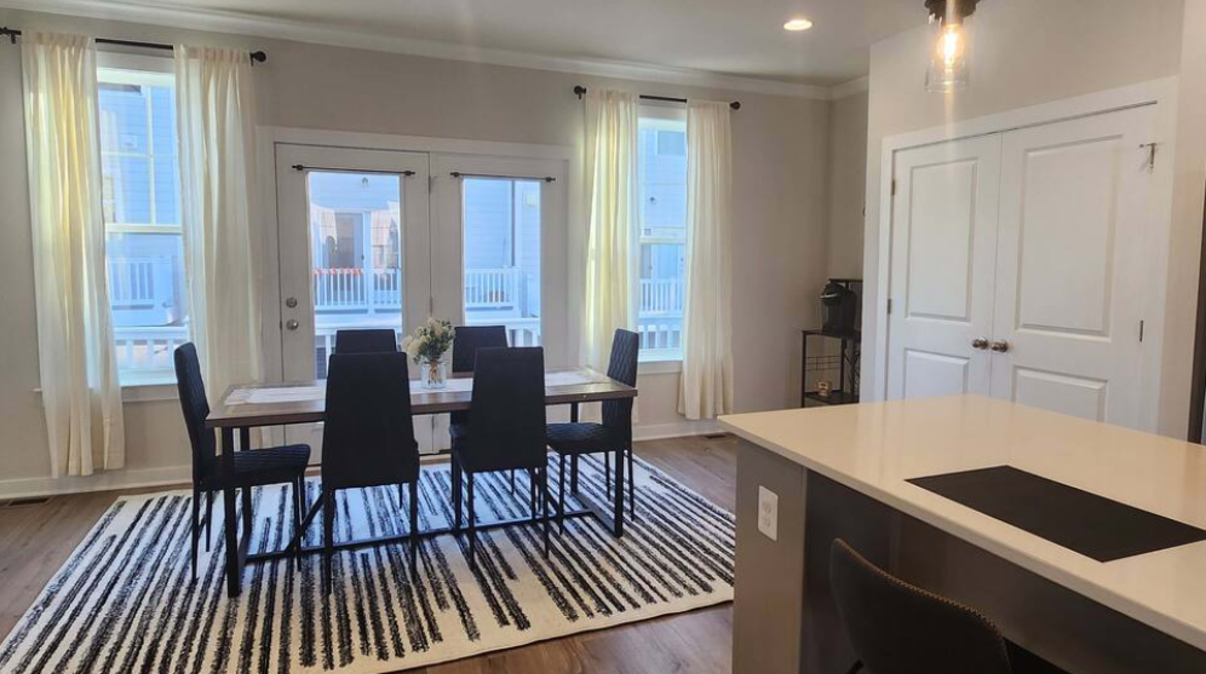 A dining area features a rectangular table surrounded by four black chairs, with a patterned rug beneath and large windows letting in natural light.