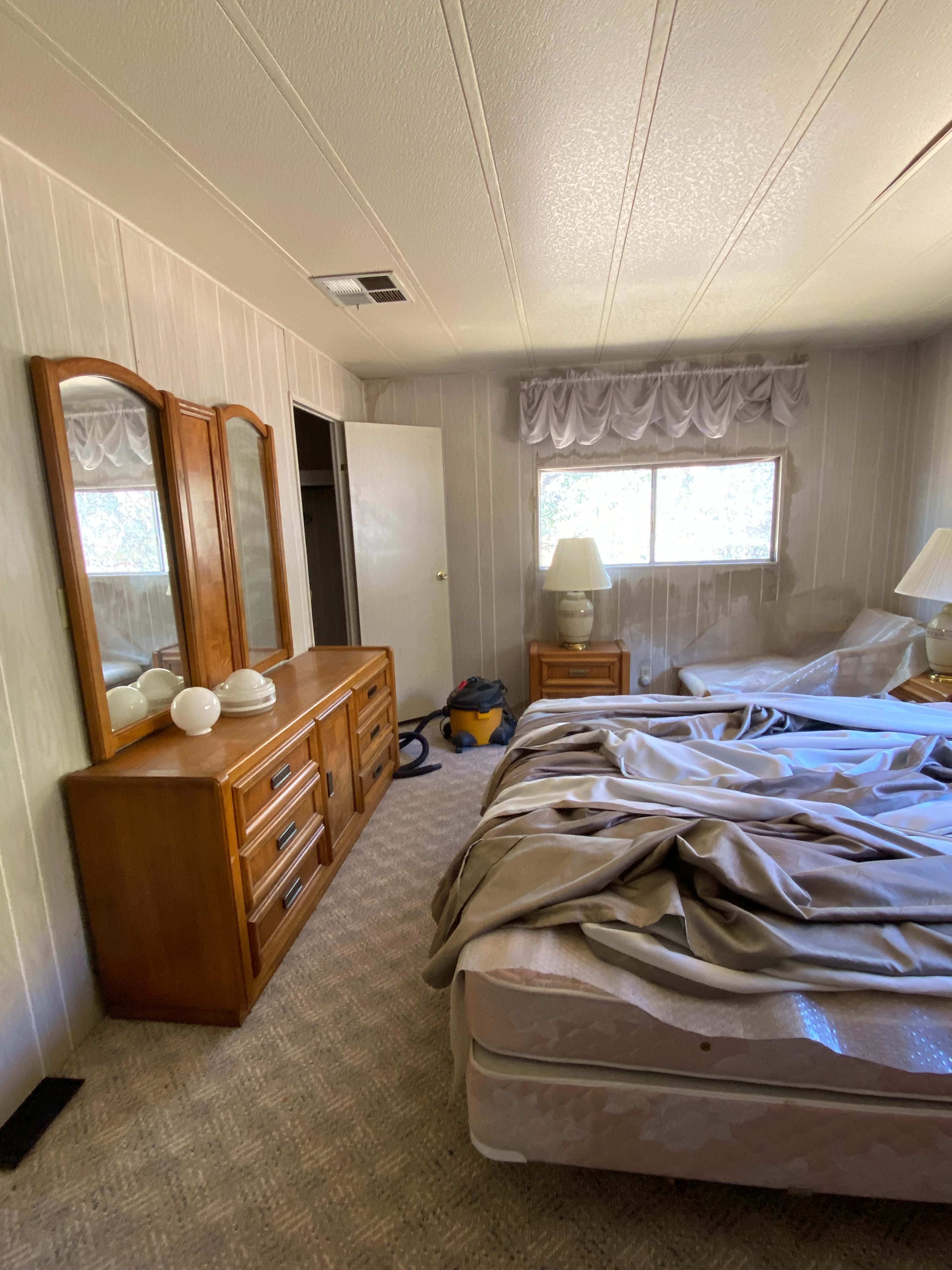 A bedroom with a bed covered in a wrinkled blanket, a wooden dresser with a mirror, and two lamps on either side of a window.