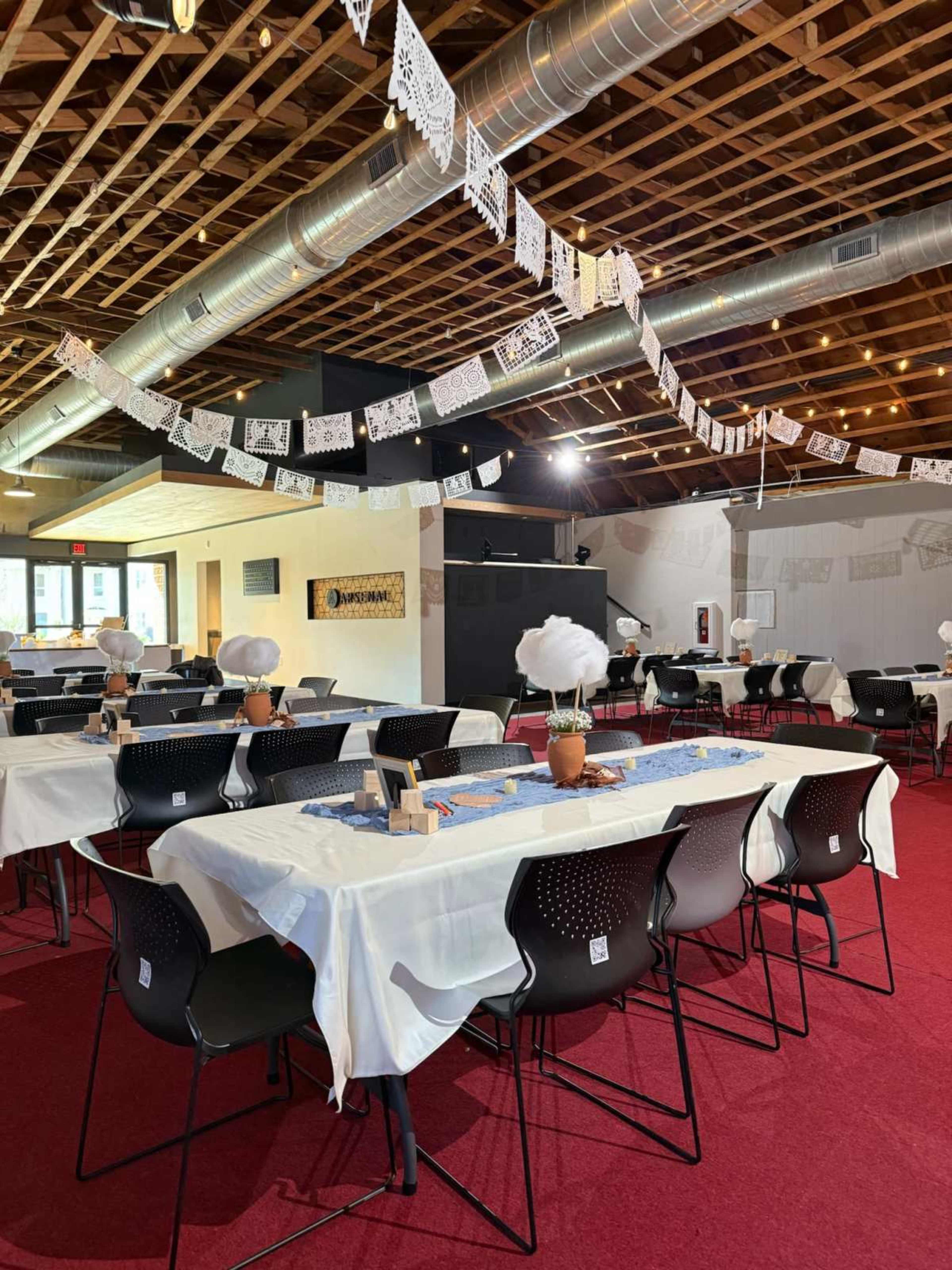 A spacious dining area features rows of tables covered with white tablecloths, black chairs, and decorative papel picado streamers hanging from the ceiling.