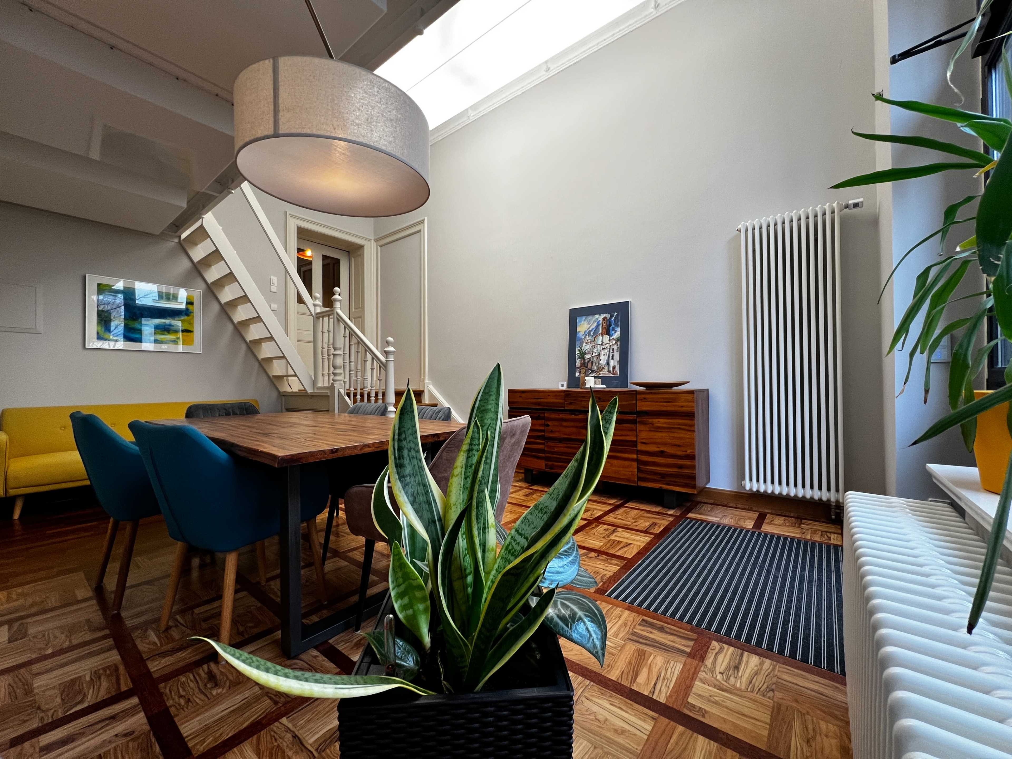 The image features a modern dining area with a wooden table surrounded by colorful chairs, a large pendant light, and a staircase leading to another level.