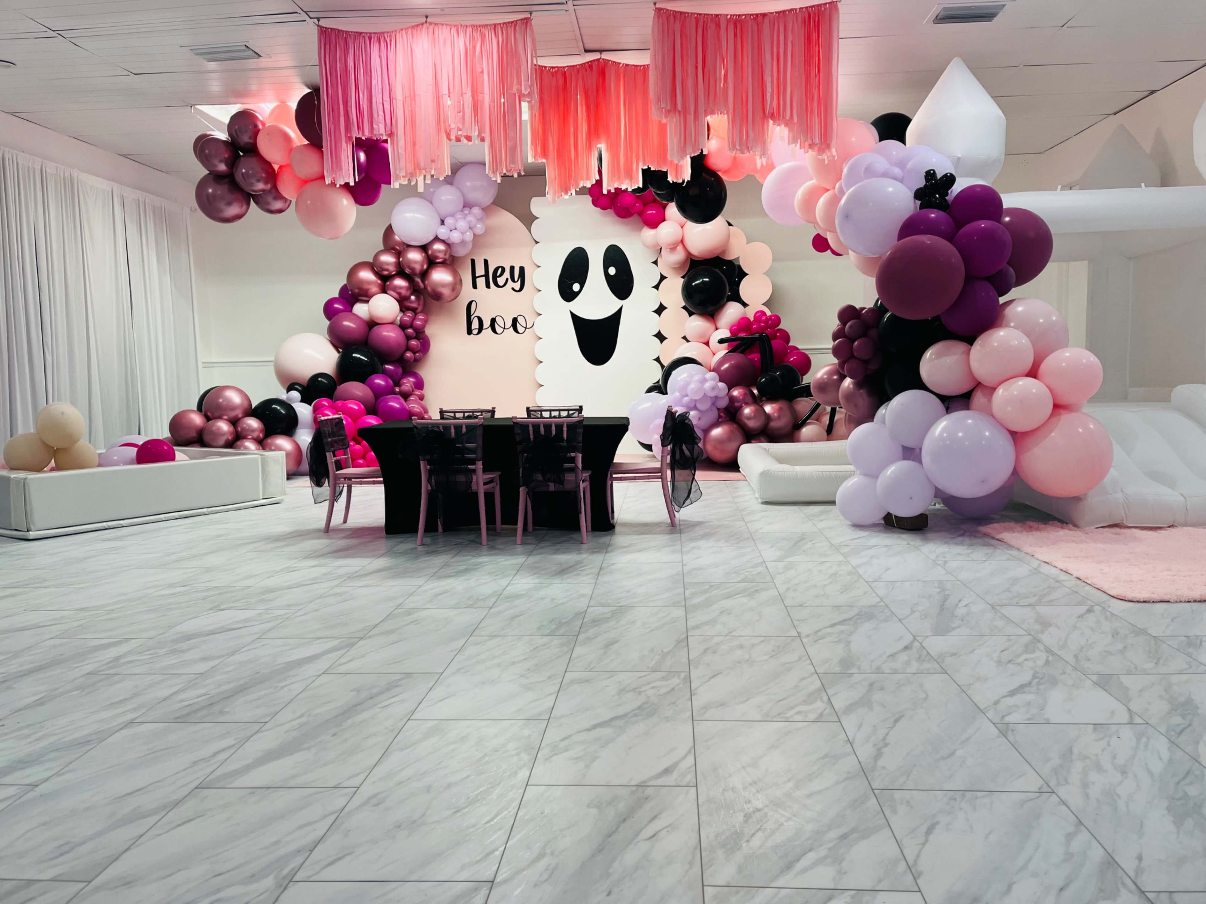 A decorated event space featuring a table set for guests, surrounded by an assortment of balloons in various colors and a wall with a ghost-themed design that reads "Hey boo."