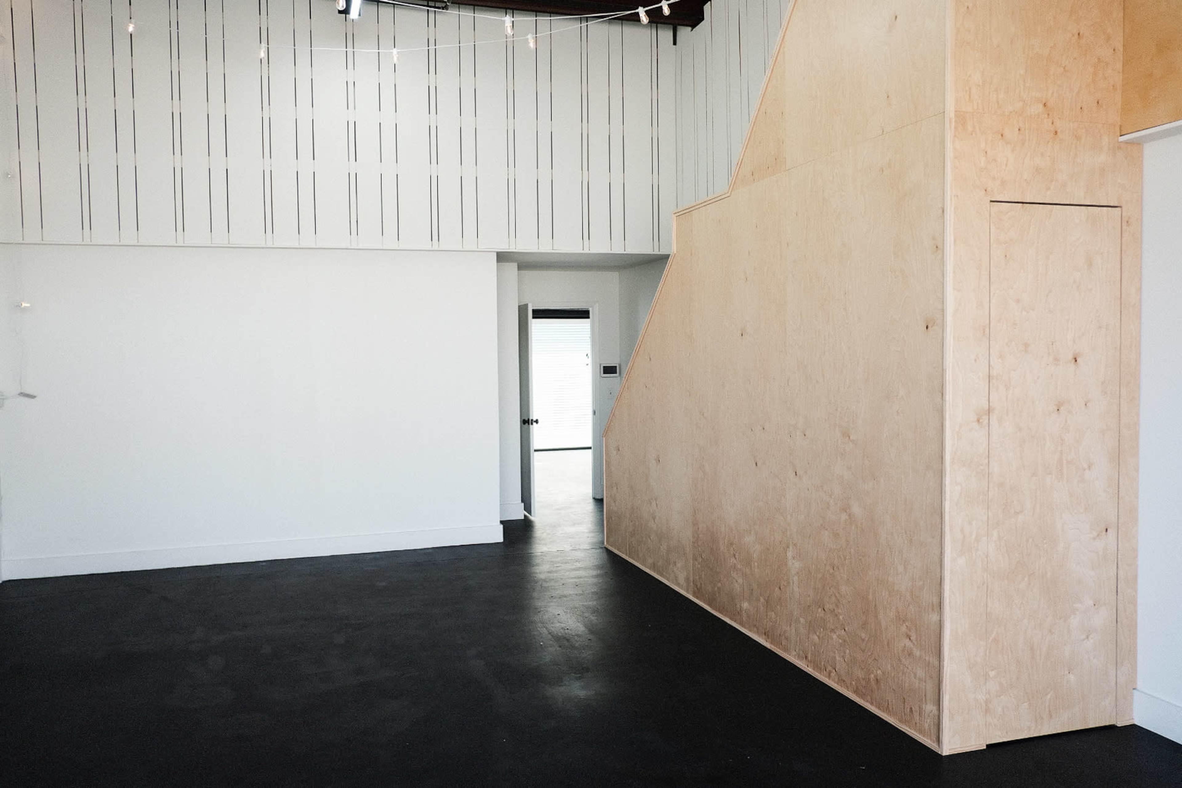 The image shows a spacious interior room with a wooden partition and stairs leading to an upper area, featuring white walls and a dark floor.