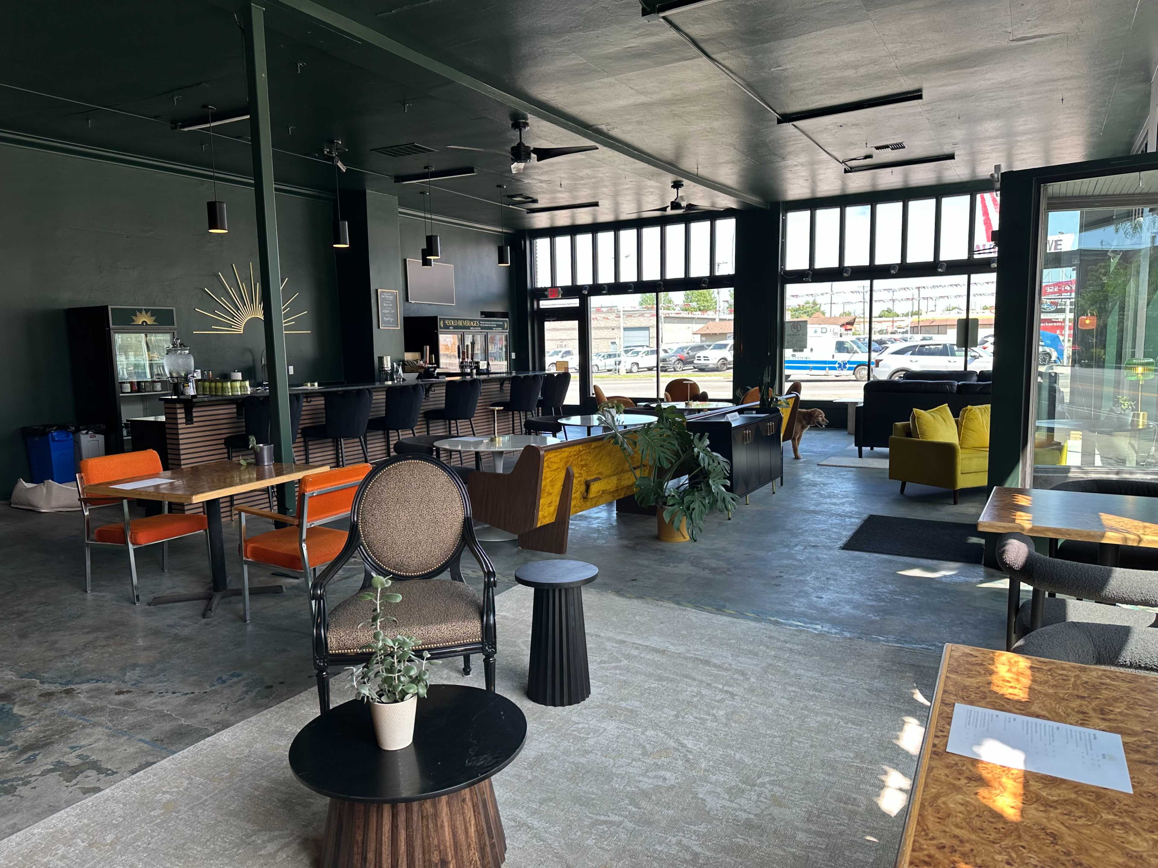 The image shows a modern café interior with various seating arrangements, including colorful chairs and tables, large windows, and a counter area in the background.