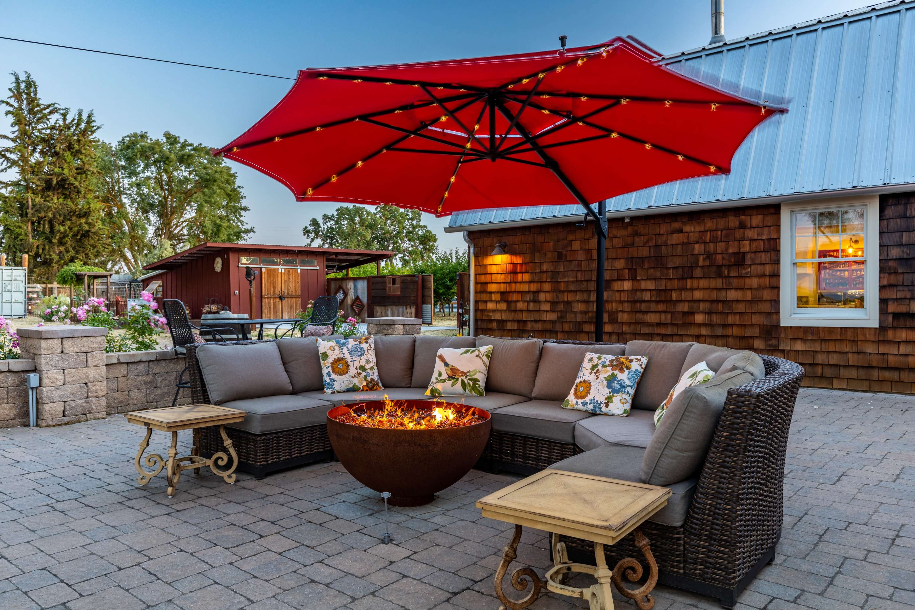 A cozy outdoor seating area features a circular fire pit surrounded by a sectional sofa and two small tables, all beneath a large red umbrella on a paved patio.