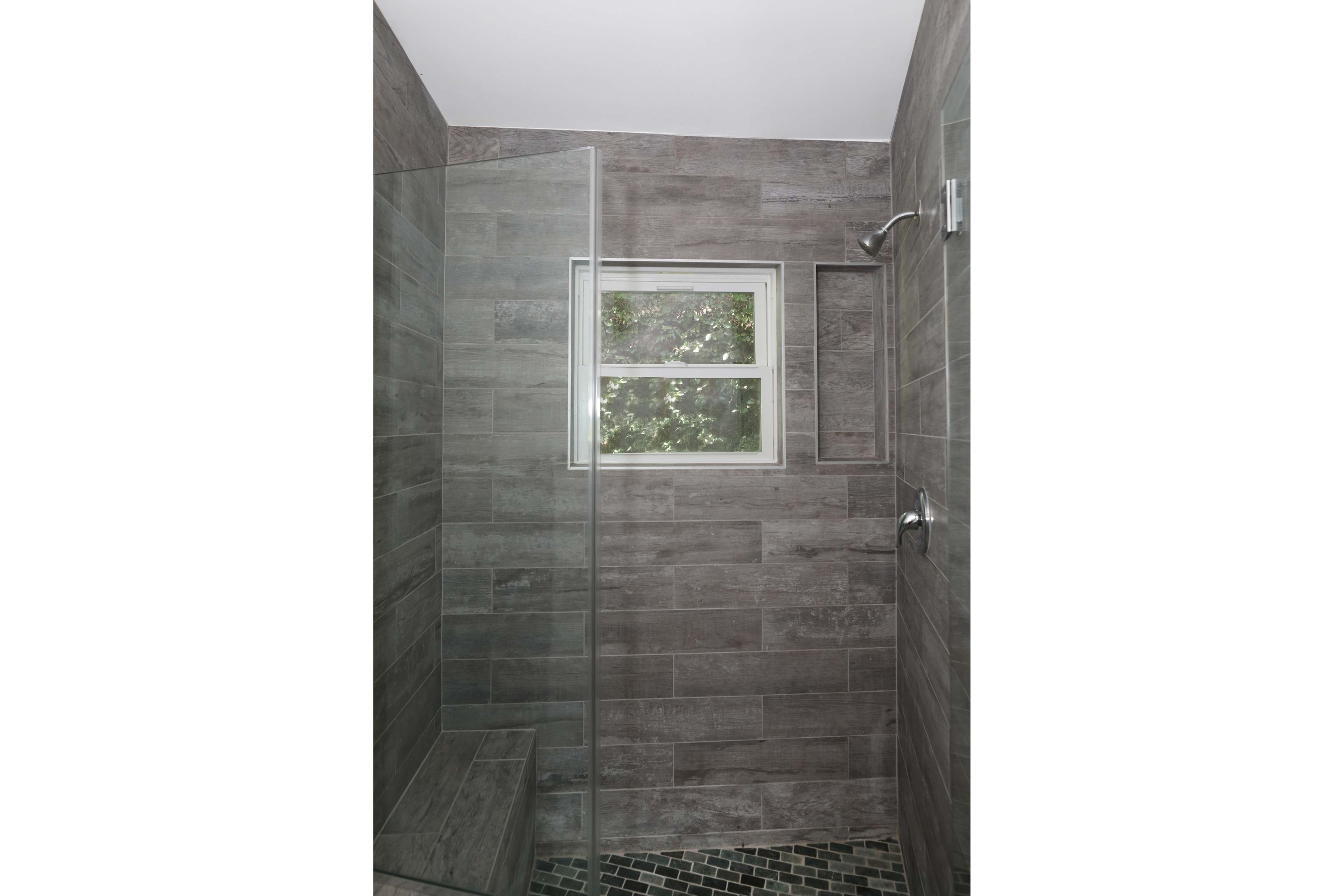 A modern walk-in shower with gray tiled walls, a glass door, and a window providing natural light.