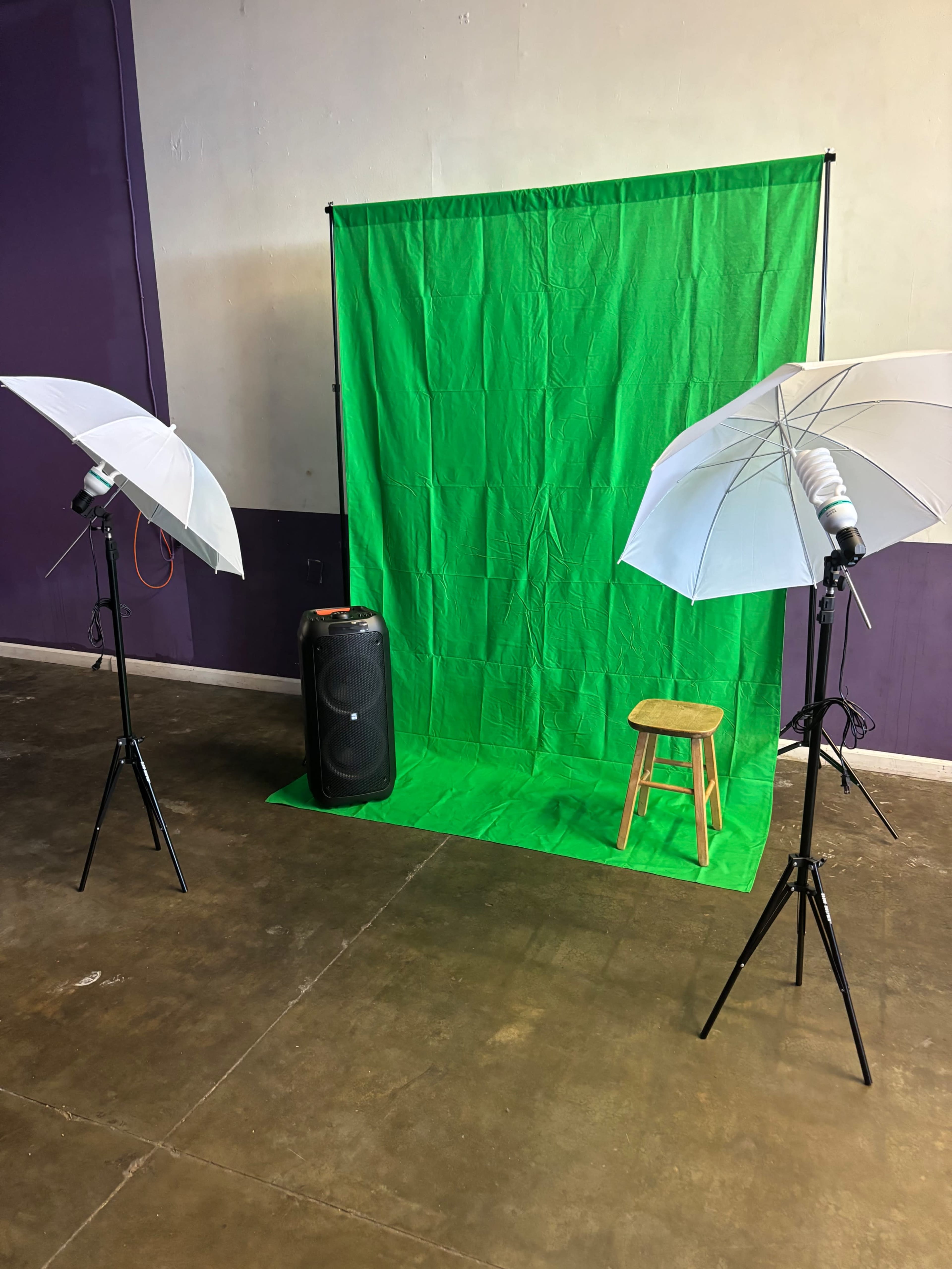 The image shows a photography setup featuring a green backdrop, two white umbrella light sources, a stool, and a speaker.