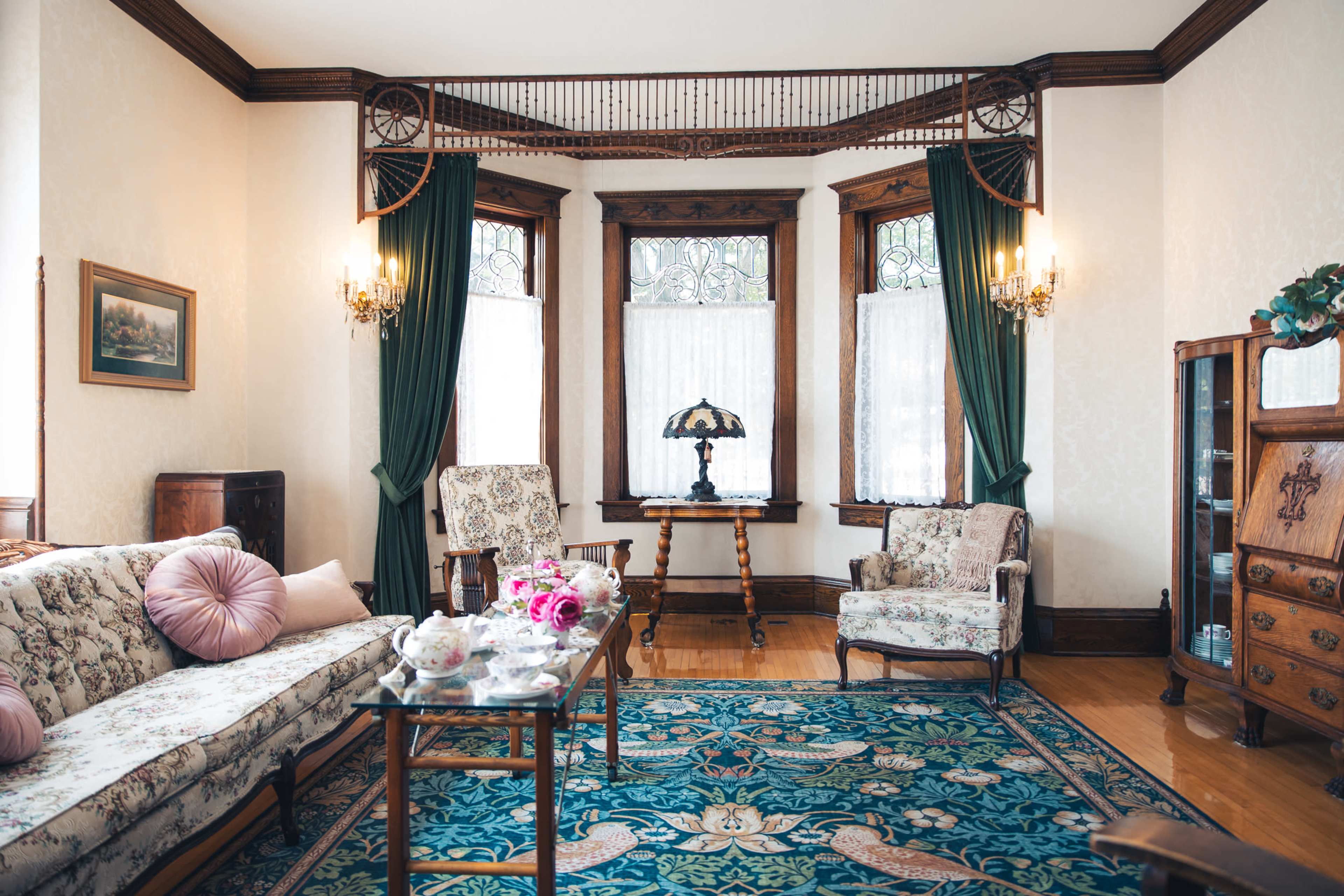 The image shows a vintage living room featuring floral-patterned furniture, elegant curtains, and a decorative rug, with large windows allowing natural light to fill the space.