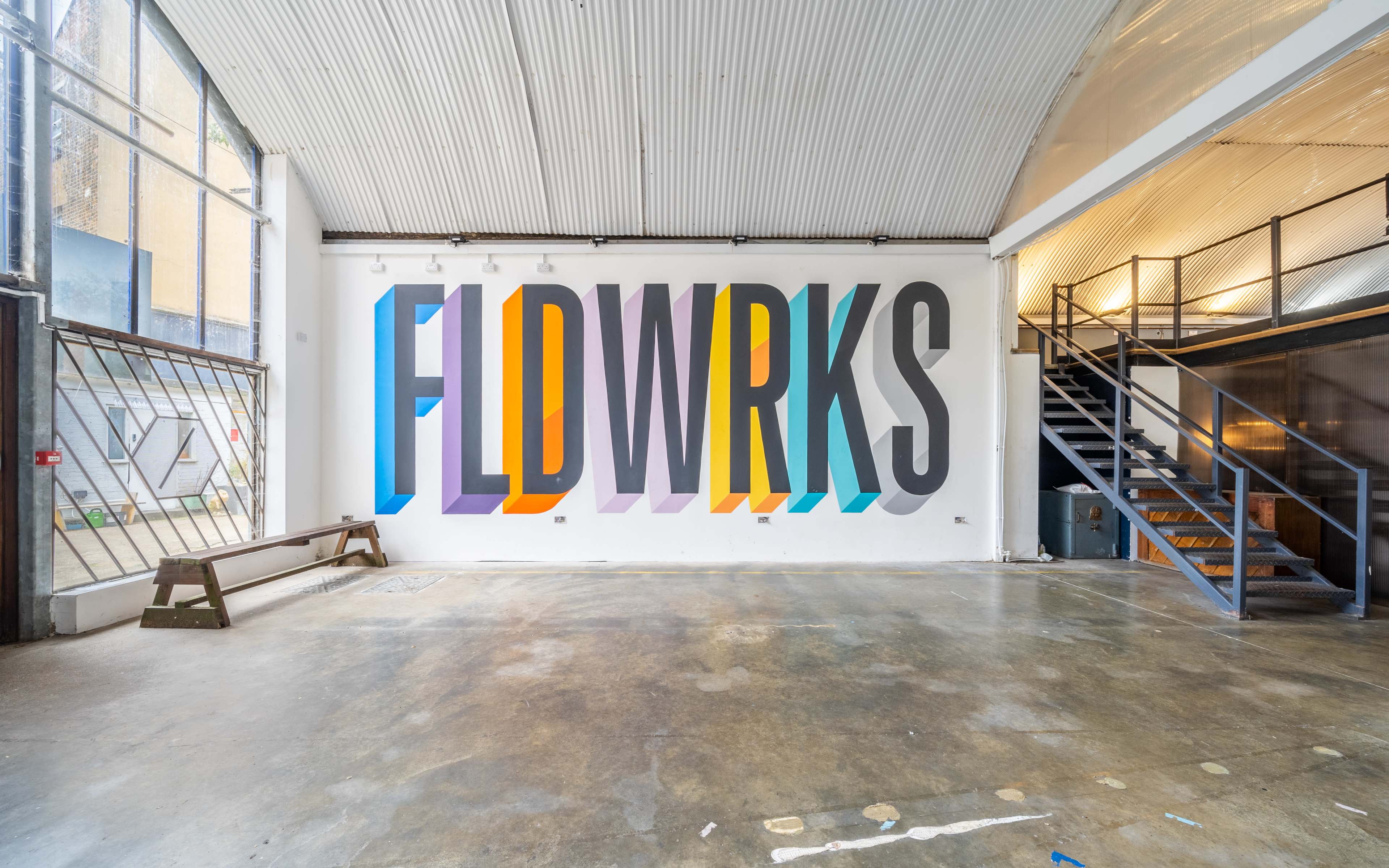 An interior space with a large mural on the wall featuring the text "FLDWRKS" in colorful, stylized letters, and a staircase in the background.