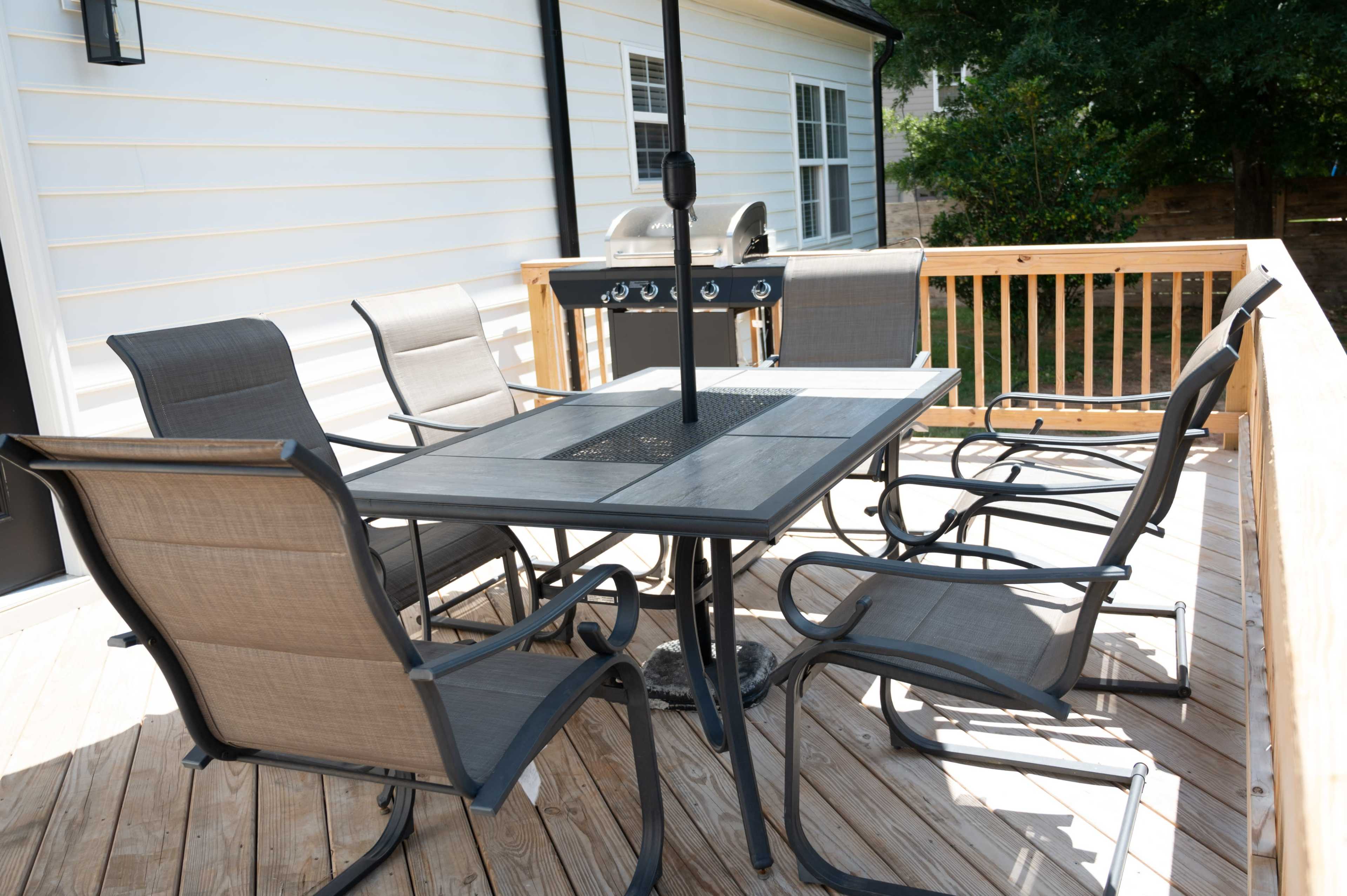 A six-seat outdoor dining table with an umbrella and a grill is positioned on a wooden deck.