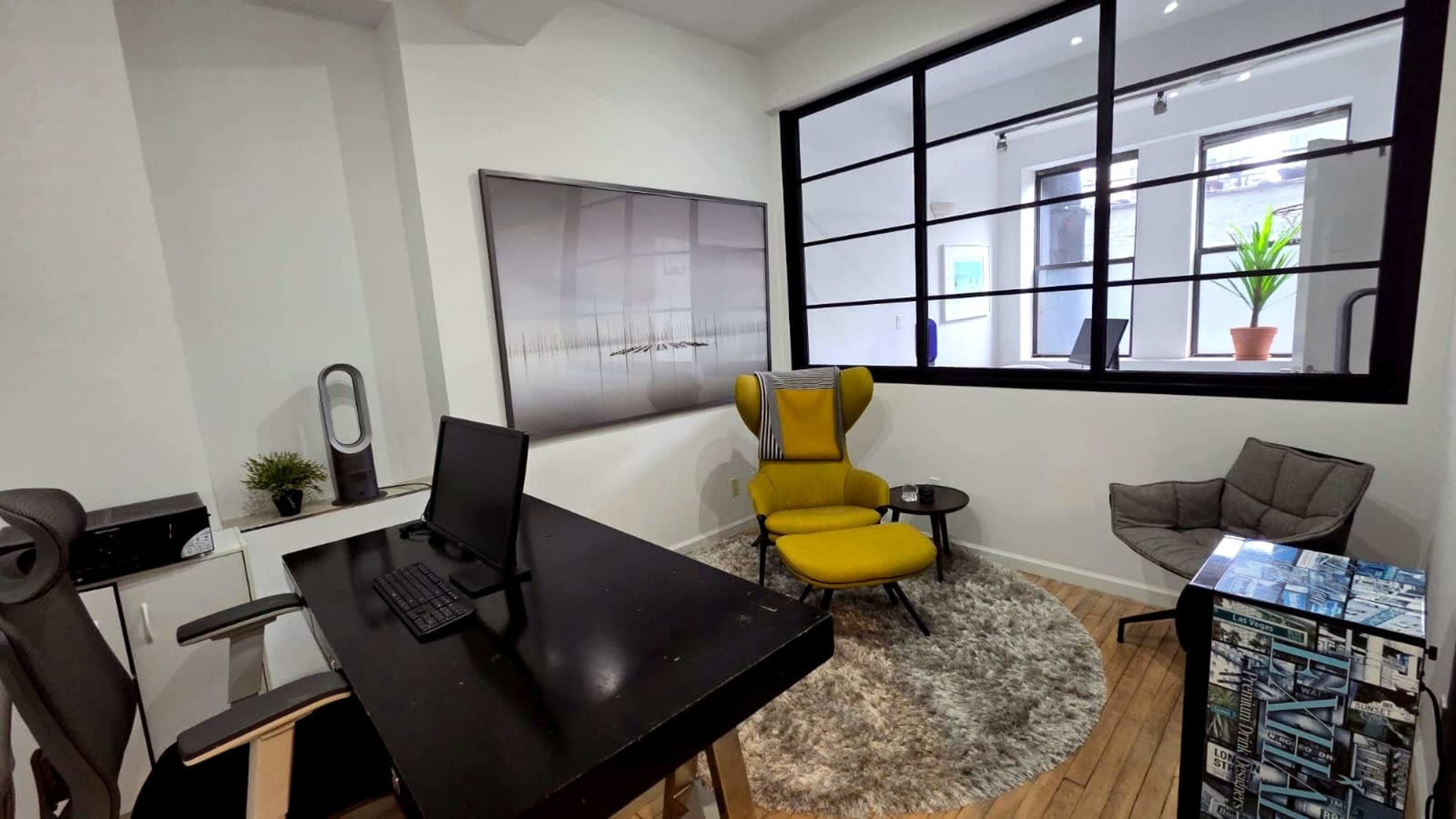 The image depicts a modern office space featuring a black desk with a computer, a yellow accent chair, a gray armchair, and large windows allowing natural light.