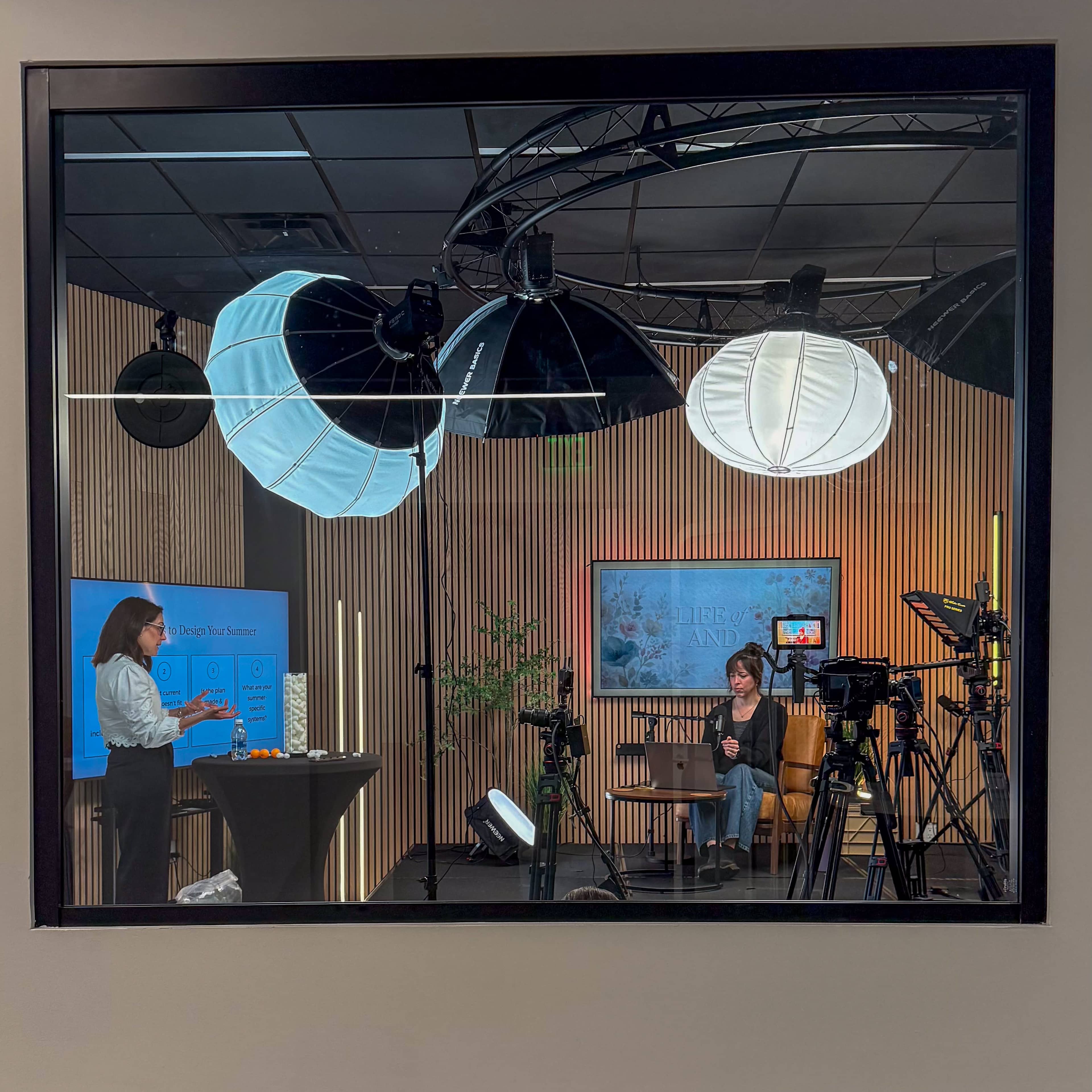 A woman stands at a table while another woman sits at a computer in a studio setting with various cameras and lights.