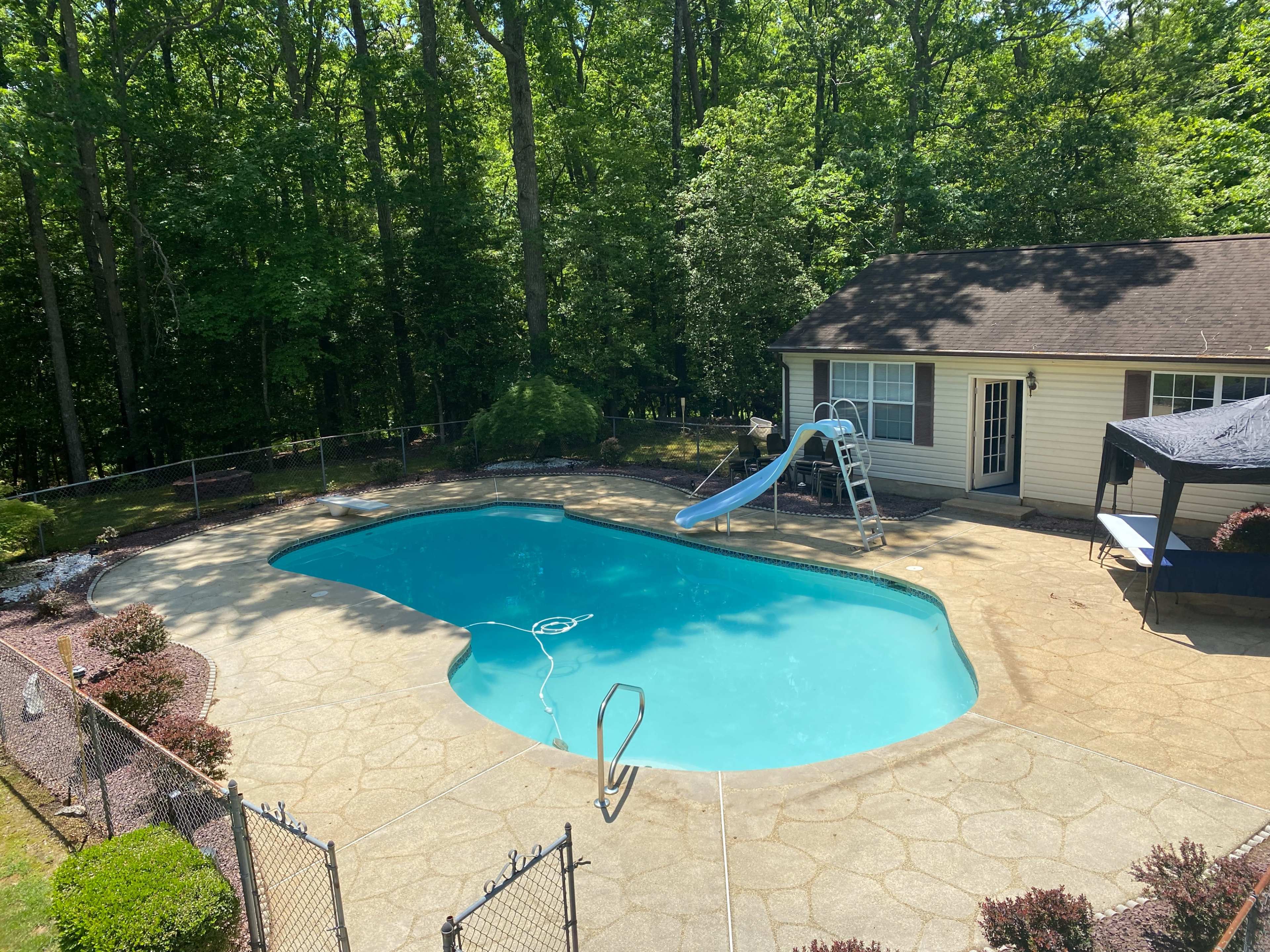 A swimming pool with a slide is surrounded by a patio and trees, adjacent to a house.