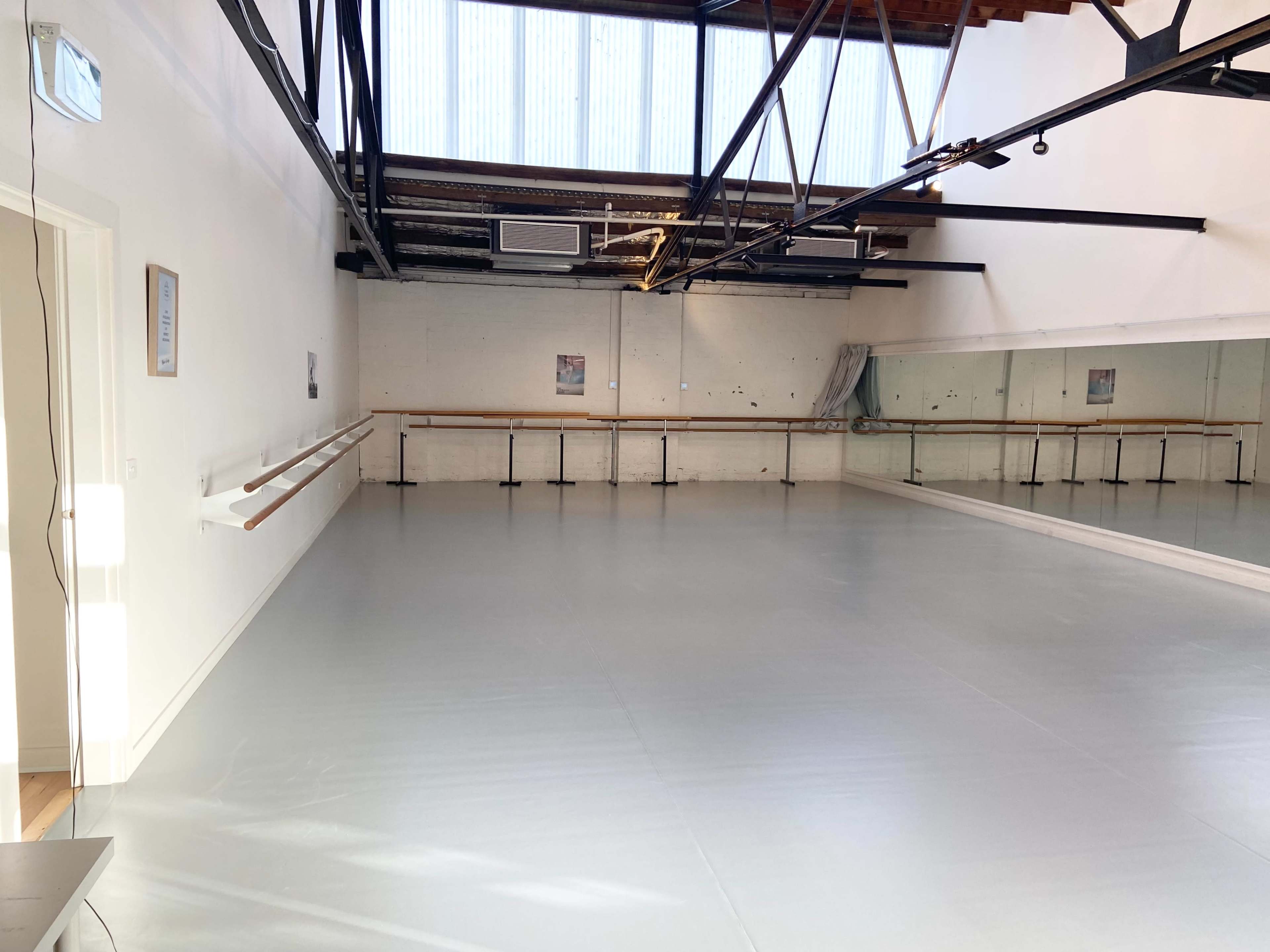 The image shows a spacious dance studio with wooden ballet bars along the mirrored wall and large windows allowing natural light to enter.