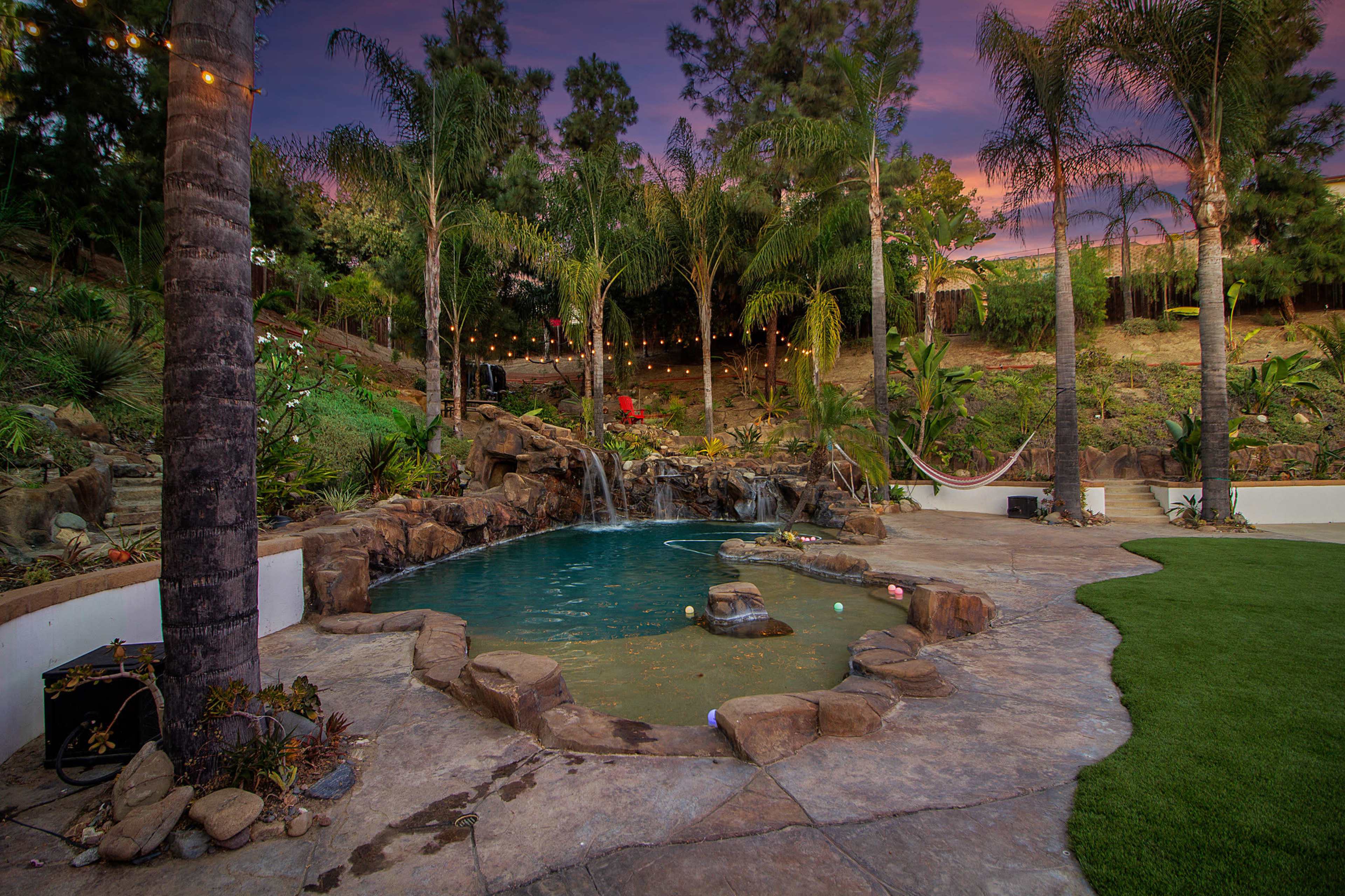 A landscaped backyard featuring a swimming pool with a rock waterfall, surrounded by palm trees and decorative lighting.