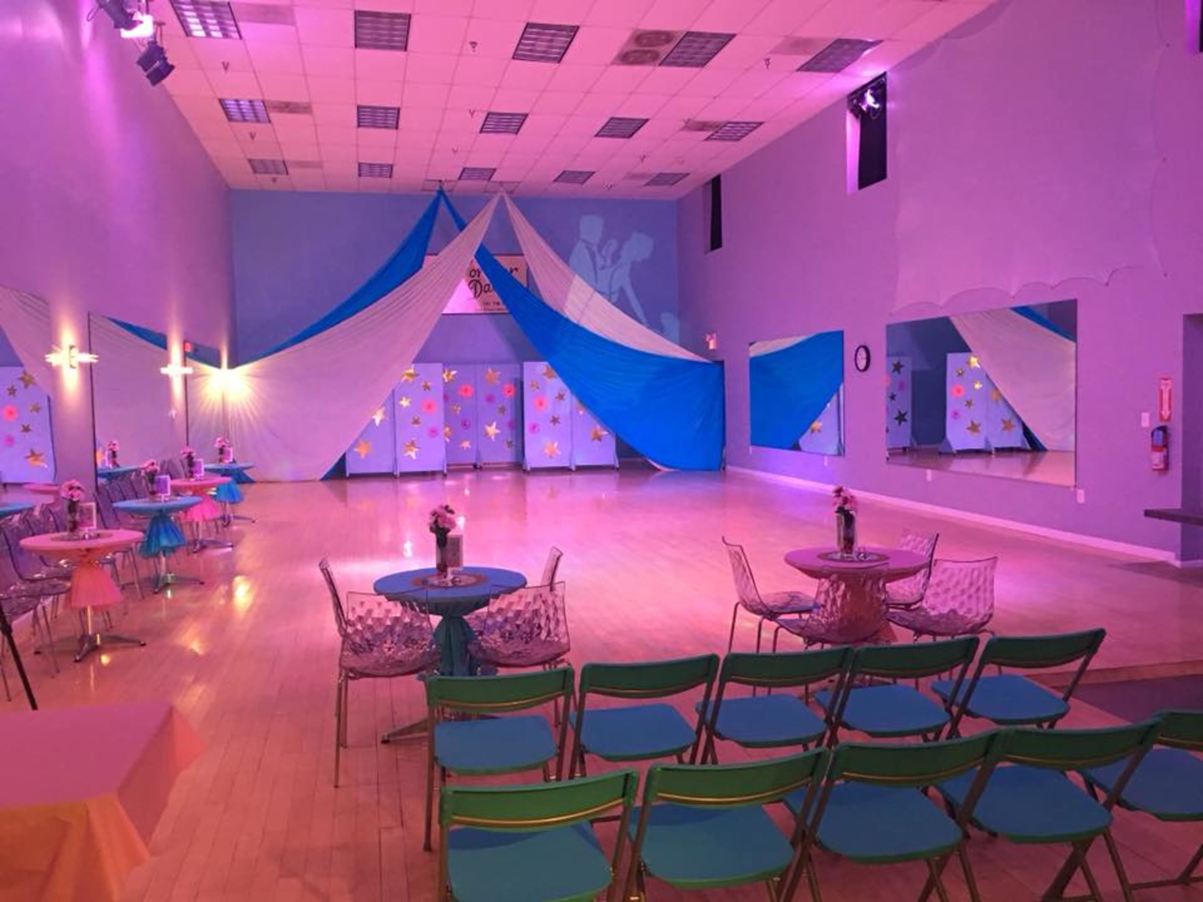 A spacious dance hall decorated with blue and white drapes, circular tables, and colorful chairs arranged for an event.