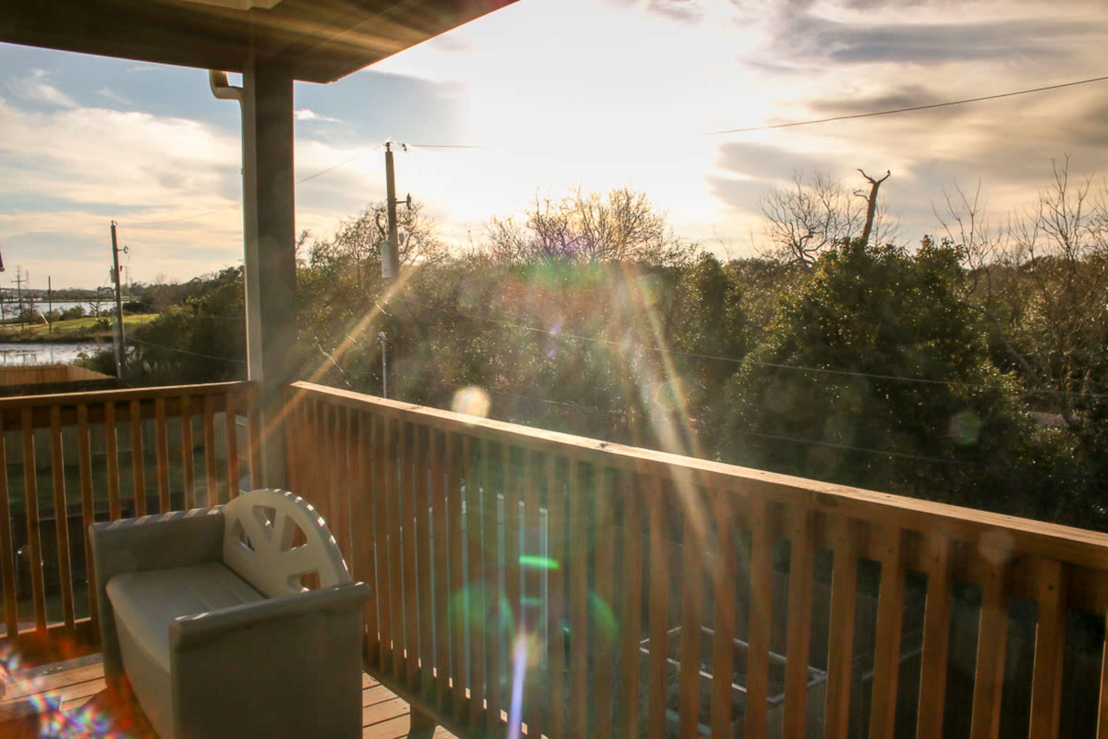 The image shows a wooden balcony with a bench, overlooking a landscape with trees and power lines under a bright sky.