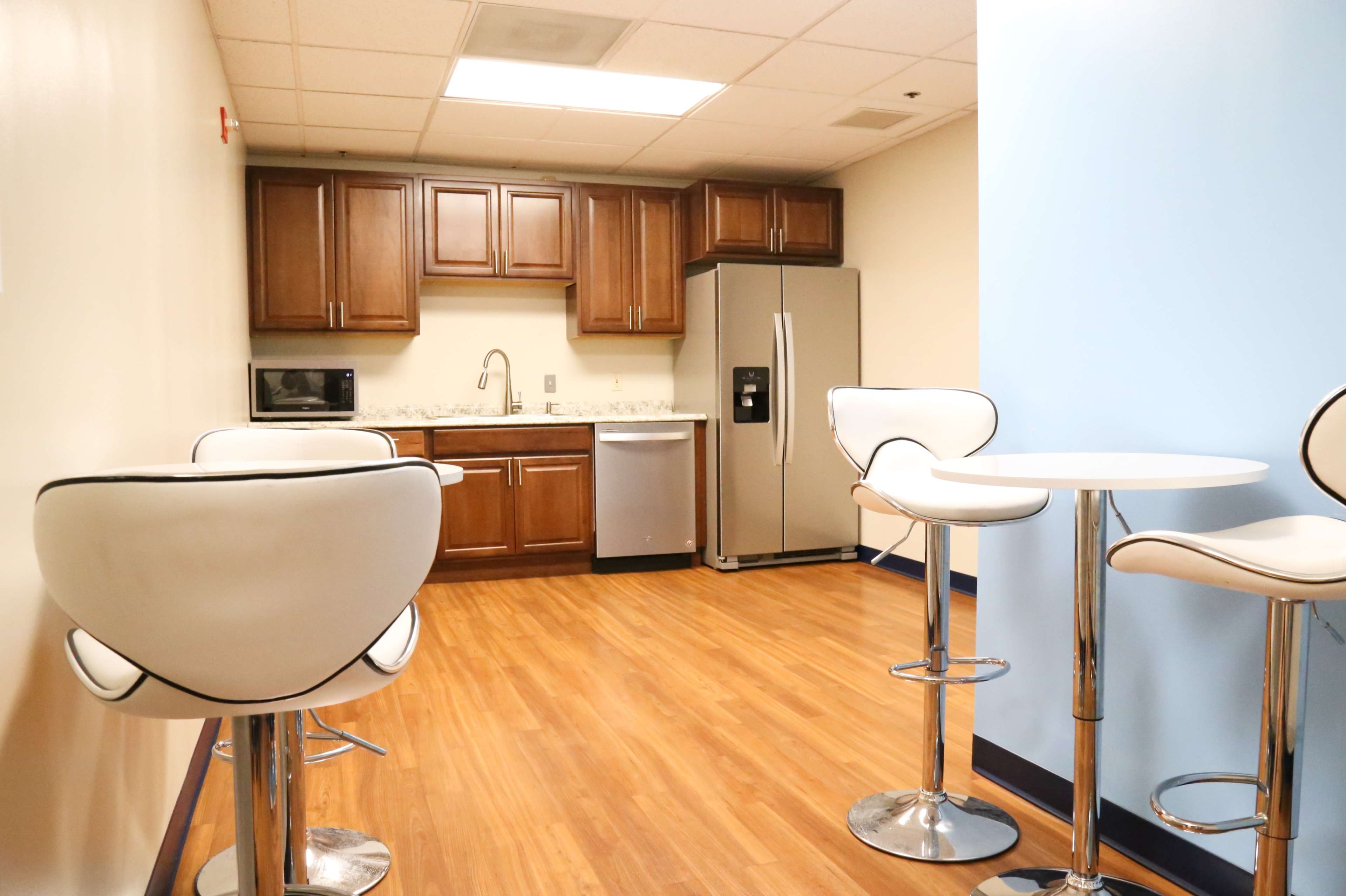 A modern kitchenette with brown cabinets, stainless steel appliances, and white bar stools around circular tables.