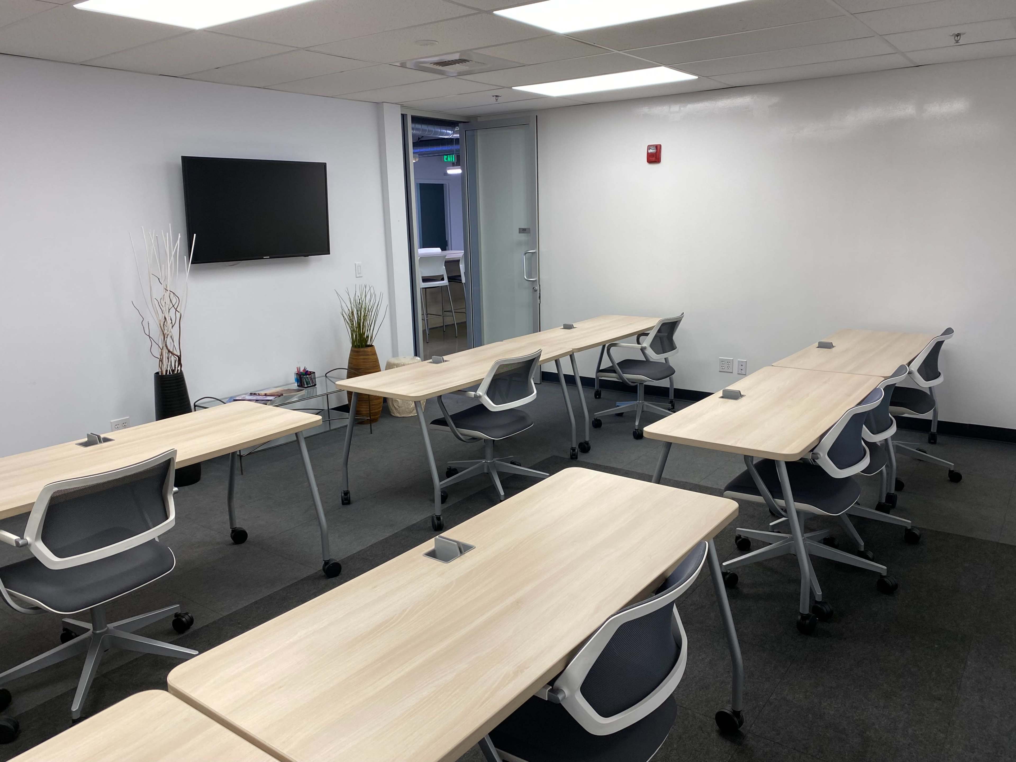 The image shows a modern classroom setup with multiple desks arranged in rows, a wall-mounted television, and a door leading to another room.