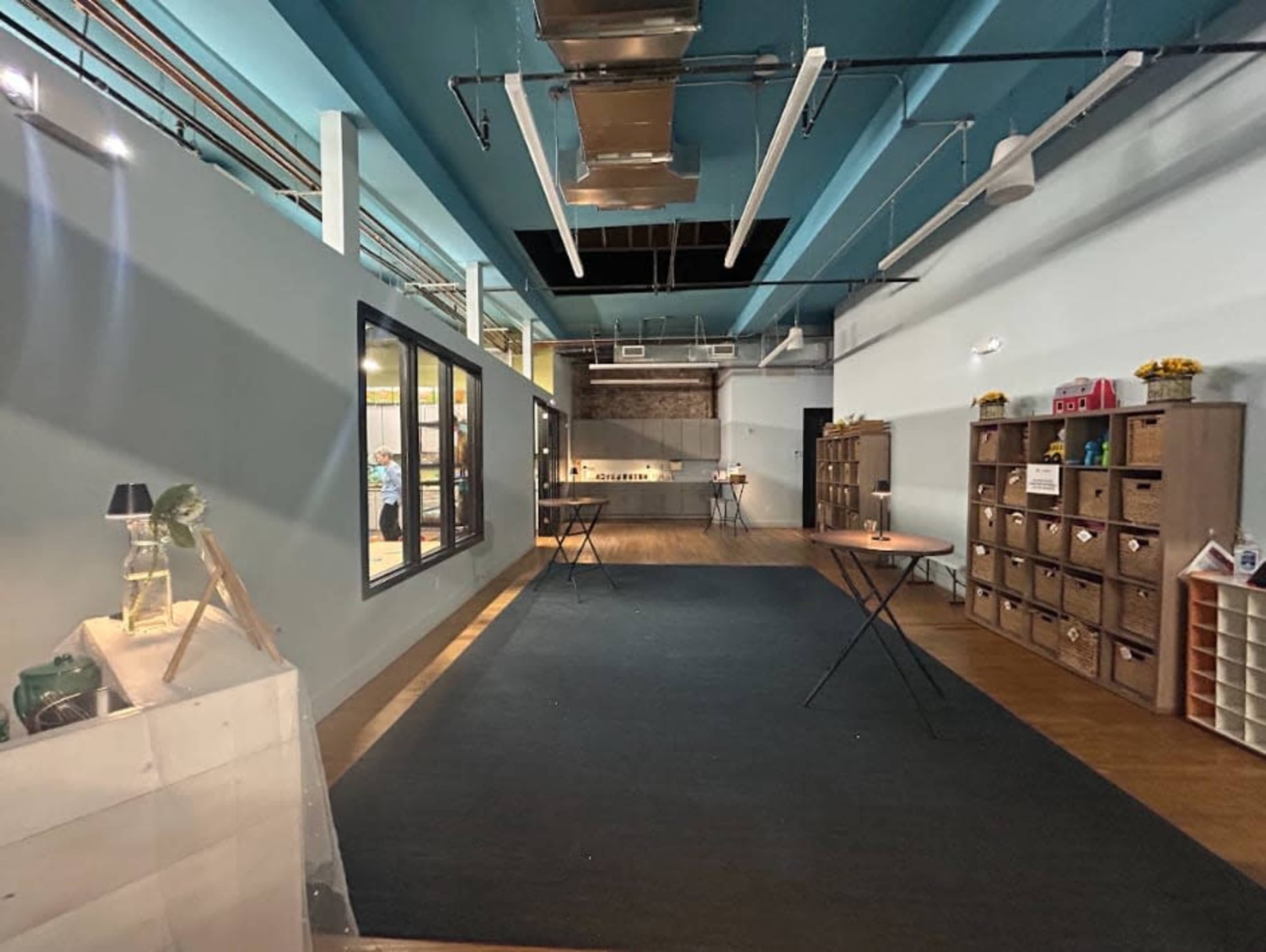 The image shows a spacious, well-lit interior with gray walls, wooden flooring, and tables set up along the sides, leading to an open area in the back.