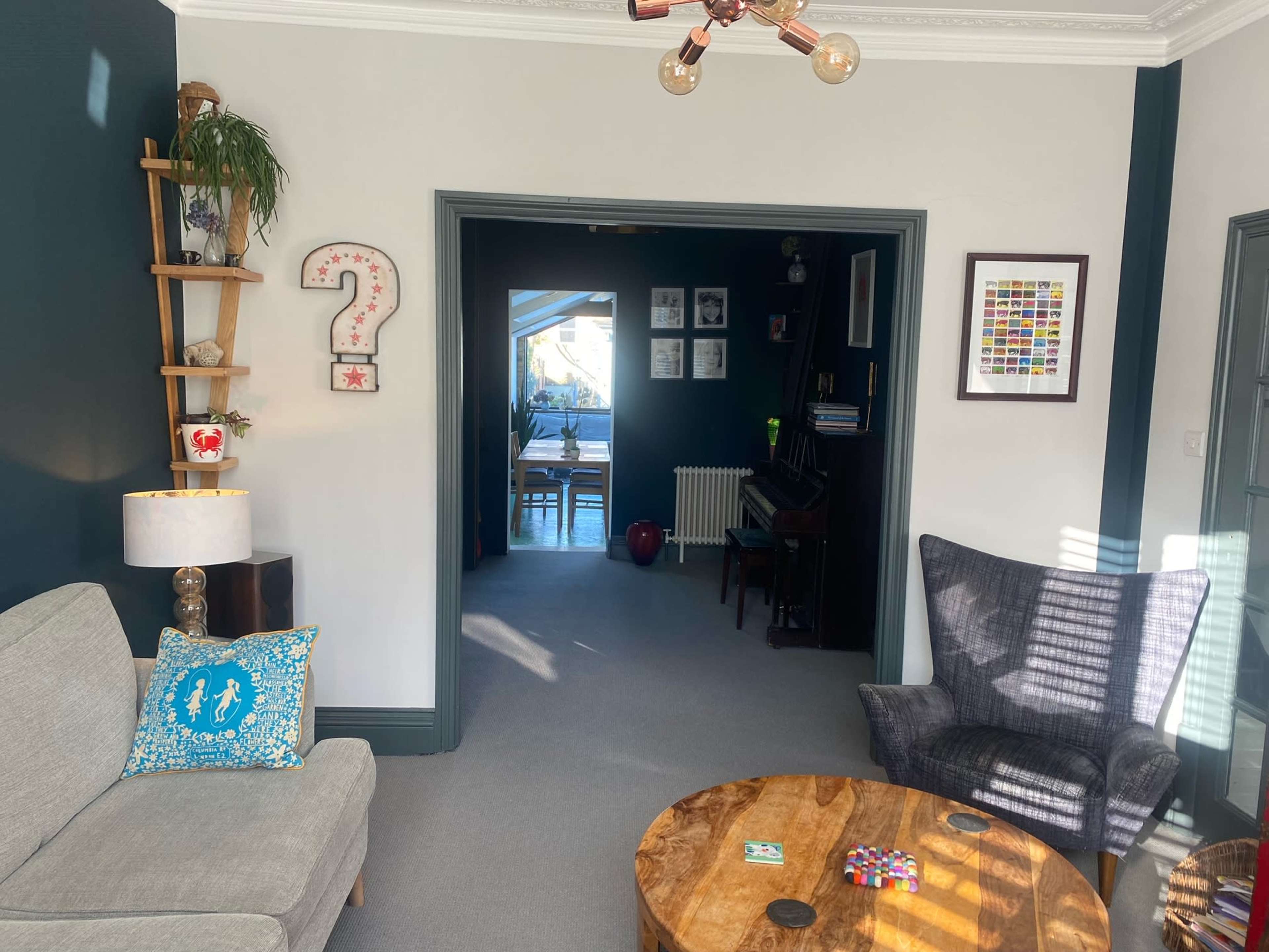 The image shows a cozy living room with a gray sofa, a wooden table, and a doorway leading to another room decorated with framed pictures.