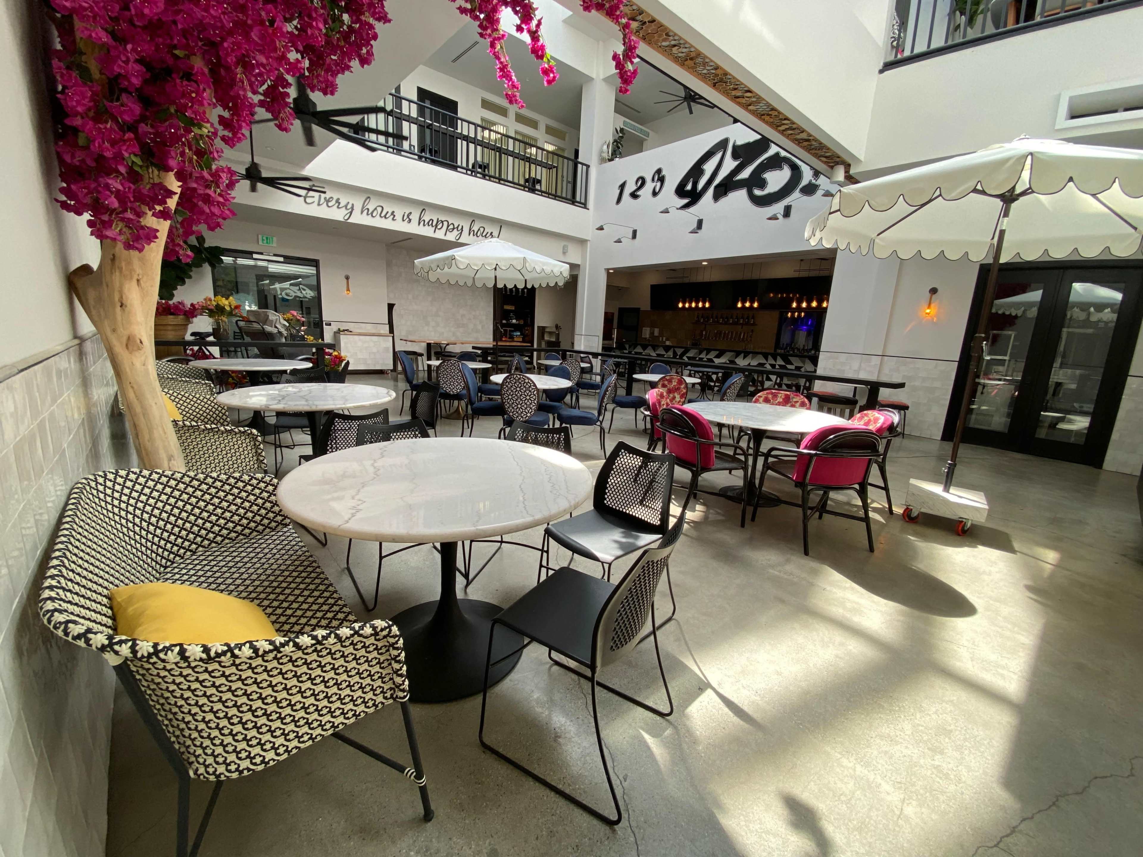 The image shows a stylish indoor dining area with several round tables surrounded by modern chairs, and decorative flowers hanging from the ceiling.