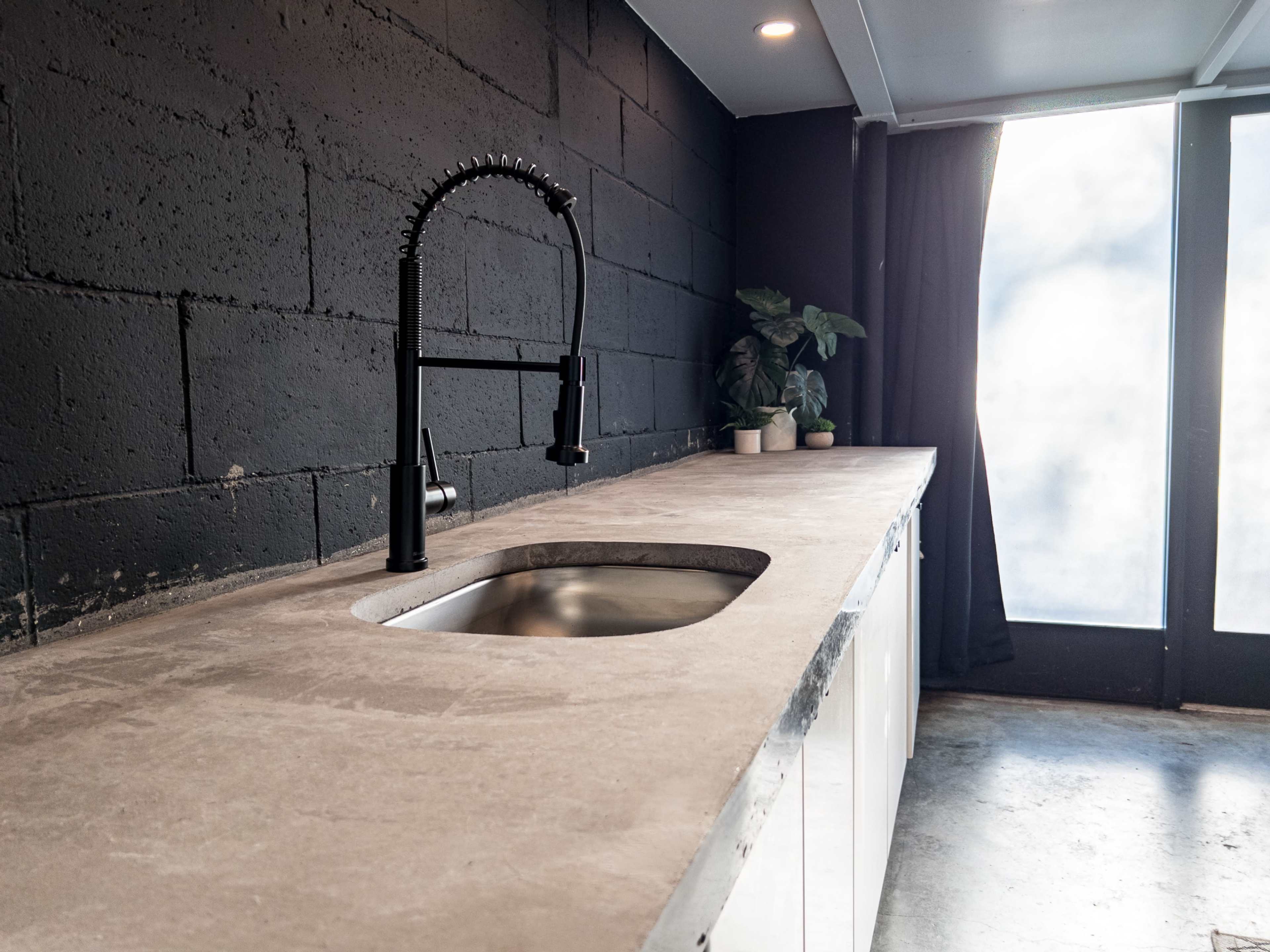 A modern kitchen features a sleek concrete countertop with a stainless steel sink and a black faucet, beside a large window covered by a curtain.