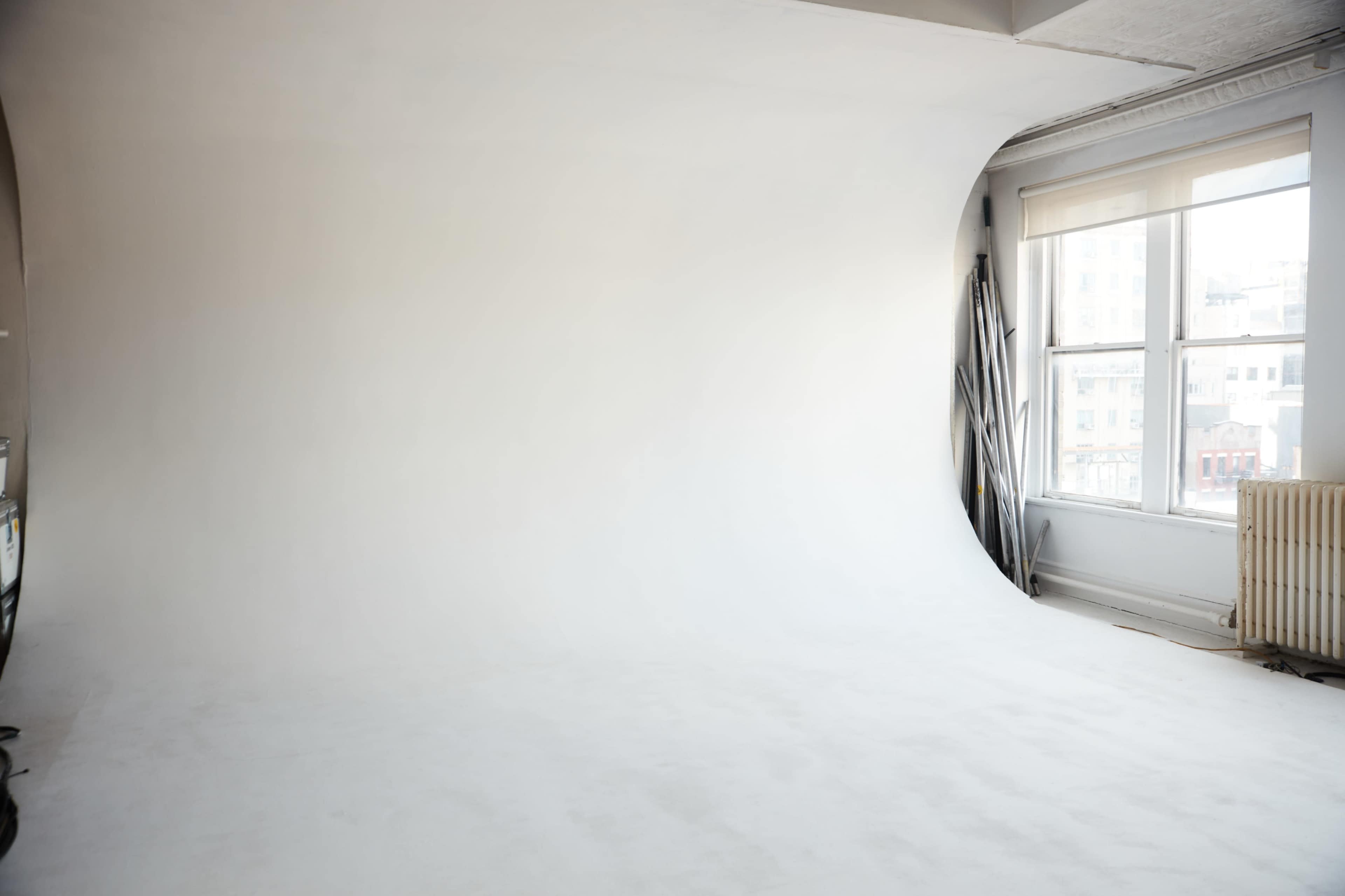 The image shows a spacious photography studio with a smooth white backdrop and large windows allowing natural light to enter.