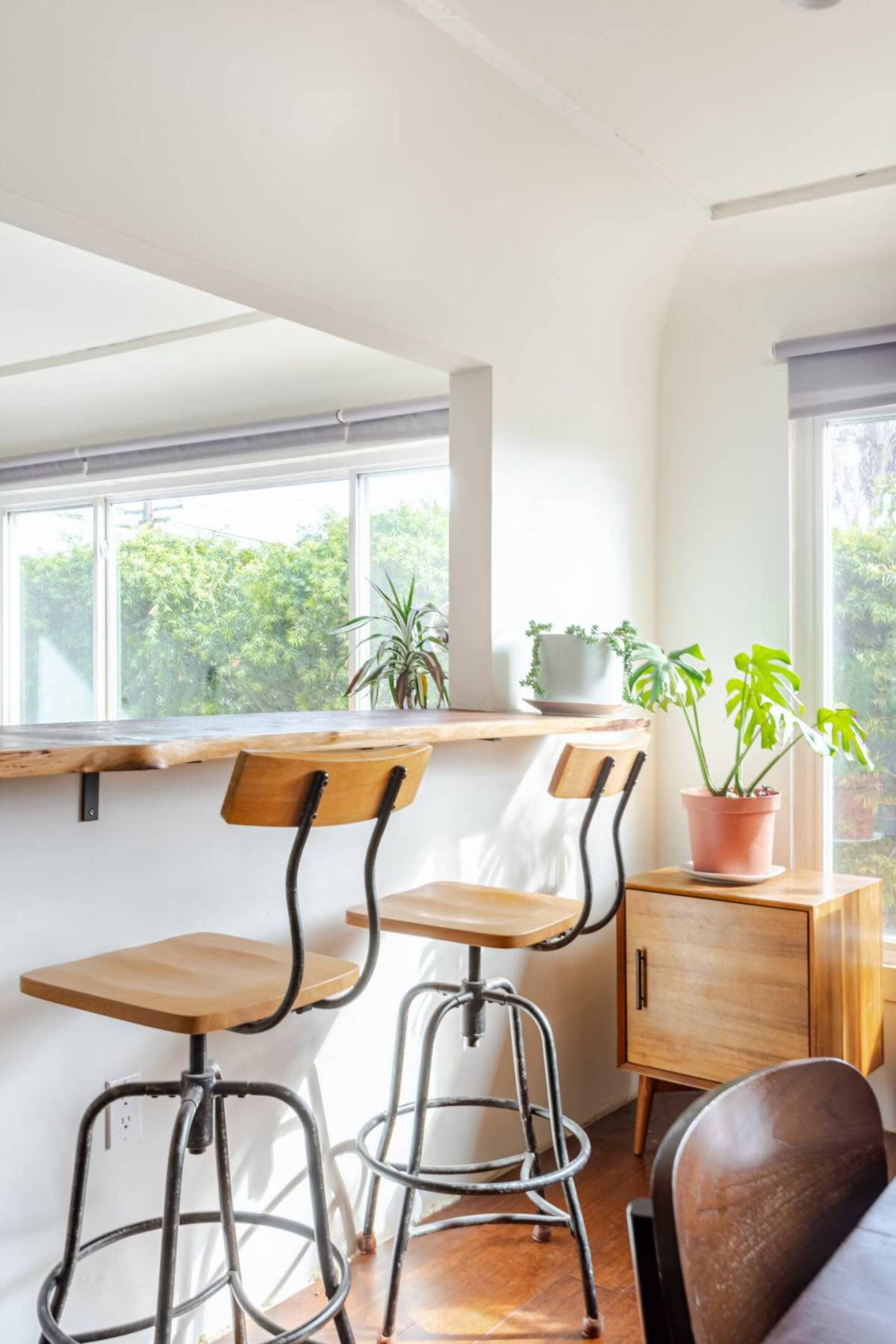 The image shows a bright, modern kitchen space with two wooden bar stools at a counter, a potted plant, and sunlight filtering through large windows.