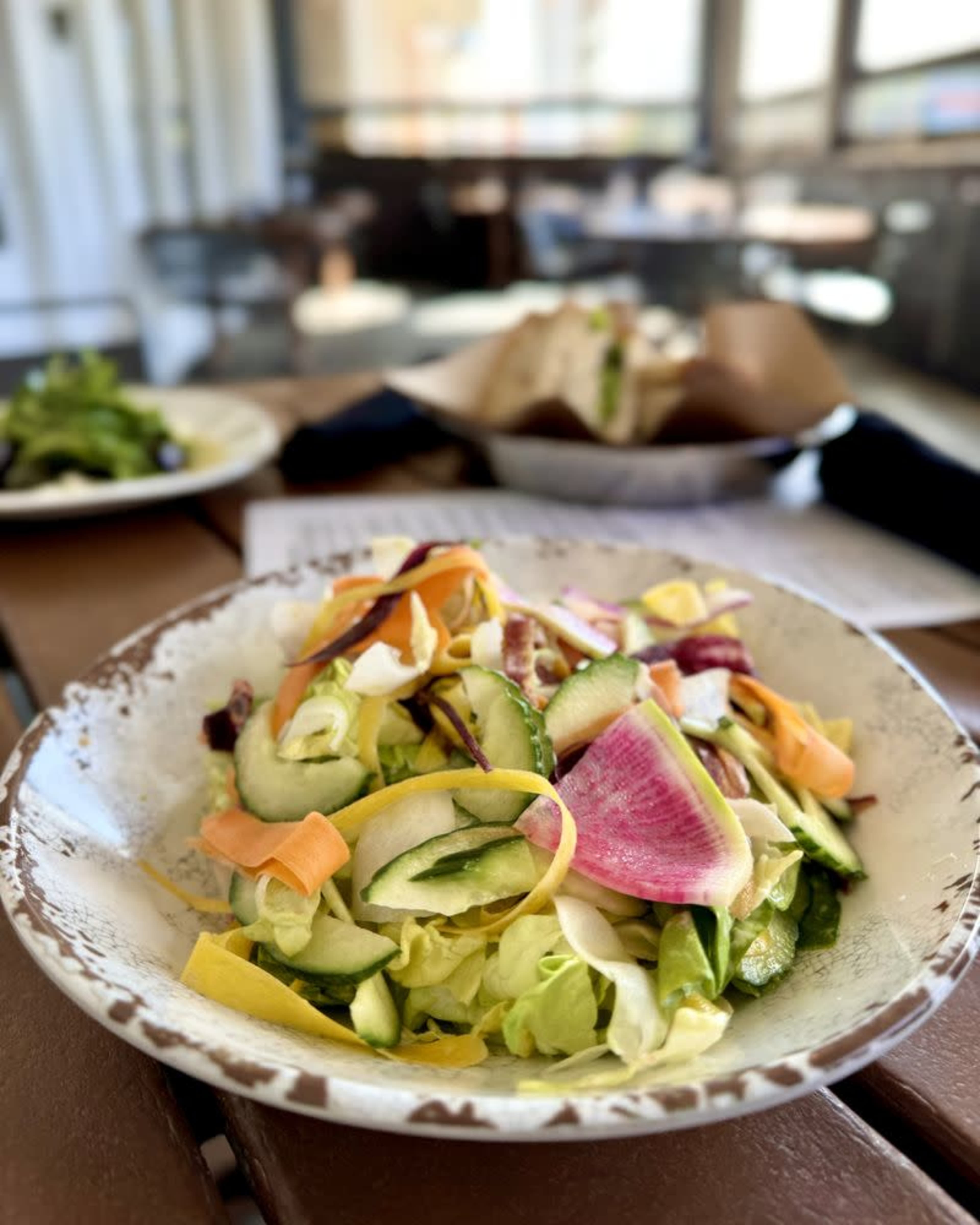 A colorful salad with mixed greens and thinly sliced vegetables is served on a patterned plate in a casual dining setting.
