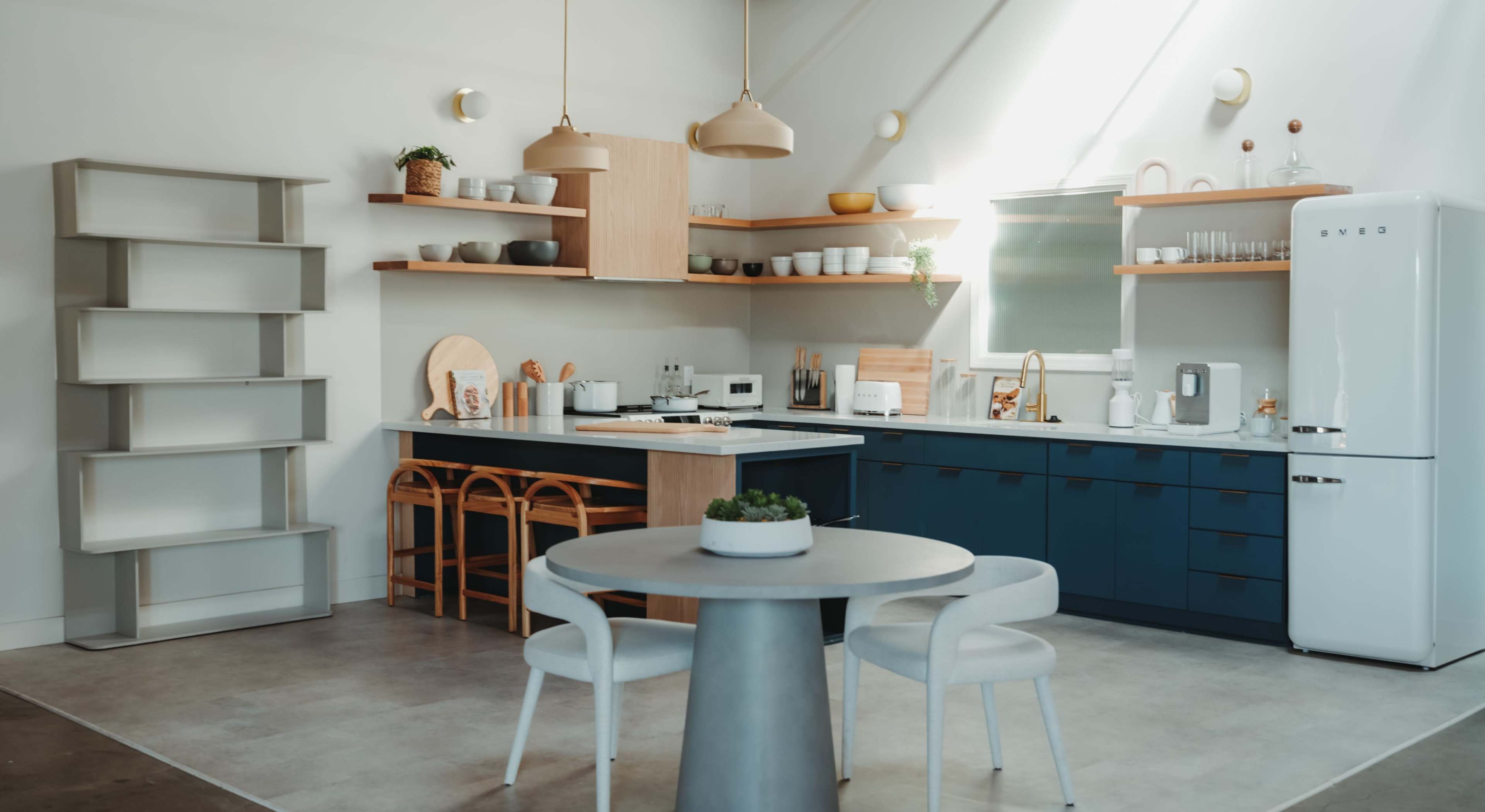 A modern kitchen features blue cabinetry, open shelving with dishware, and a circular dining table with white chairs.