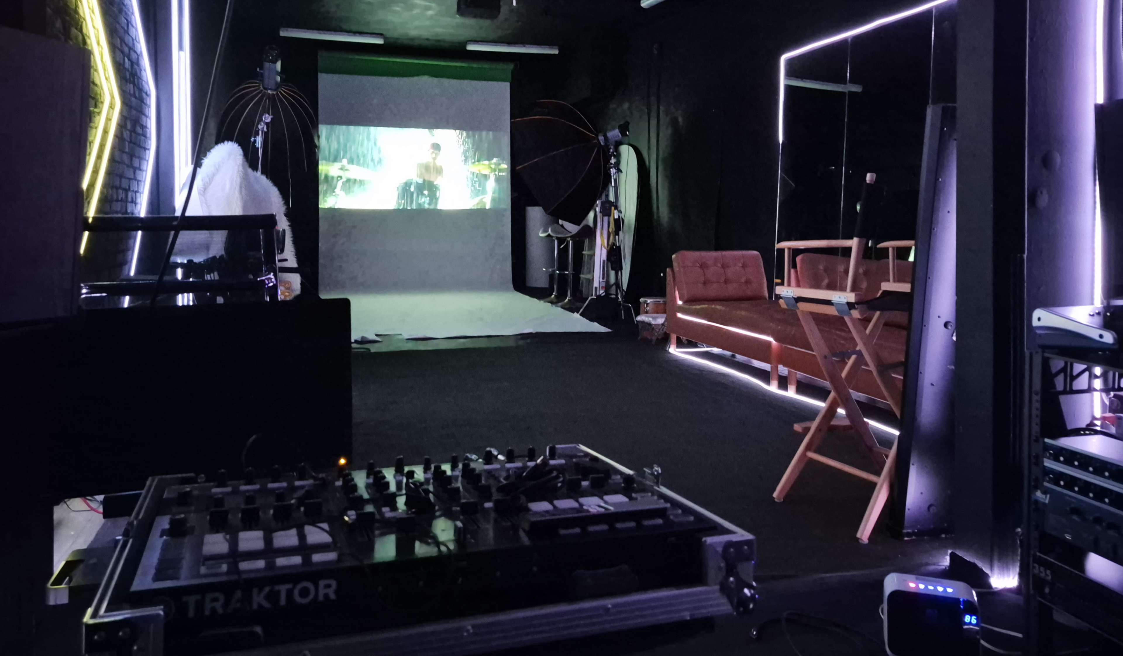 The image shows a dimly lit studio with a projection screen displaying an image, a DJ controller on a table, and a wooden chair positioned nearby.