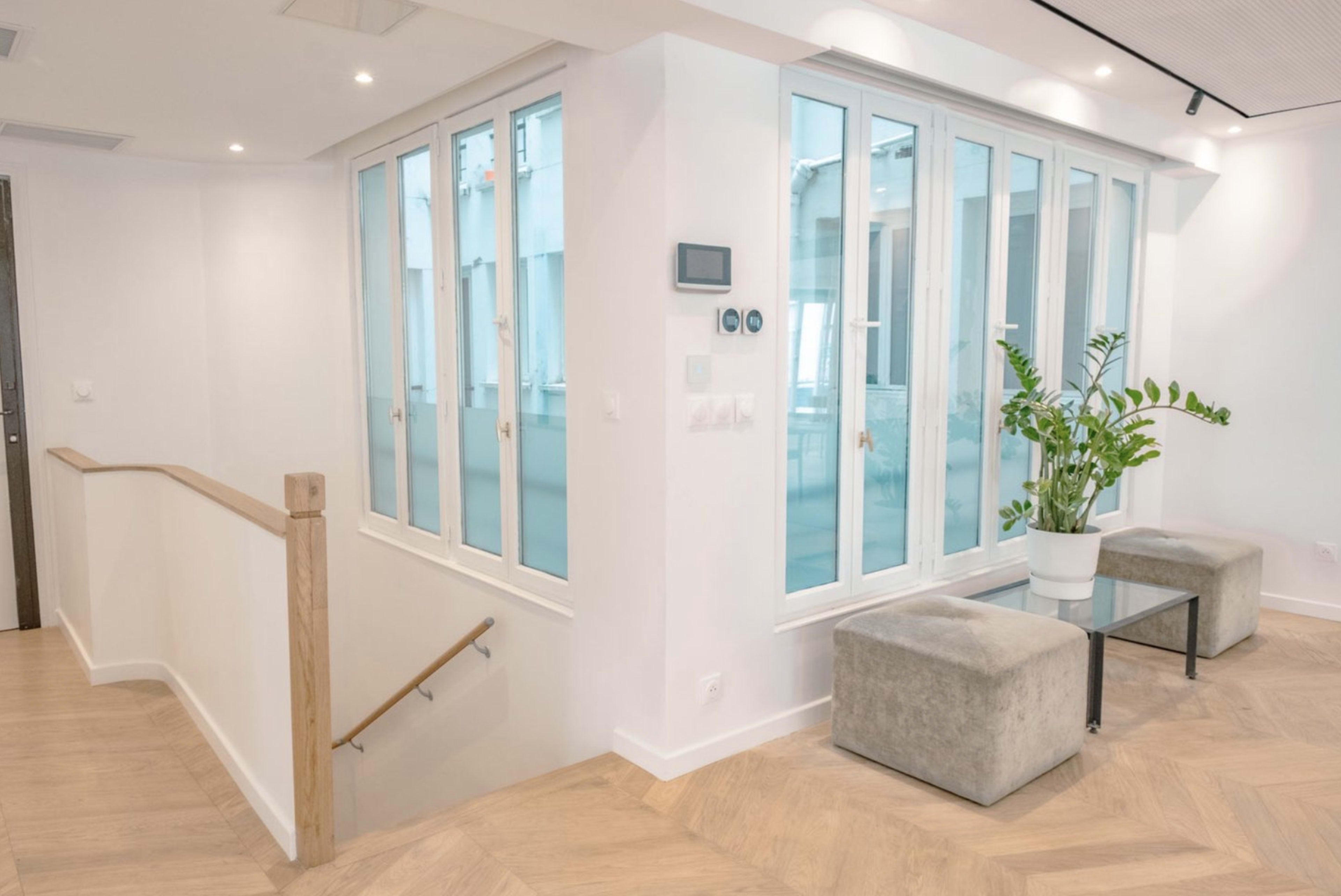 The image shows a modern interior space with a small seating area, large windows, and a potted plant.