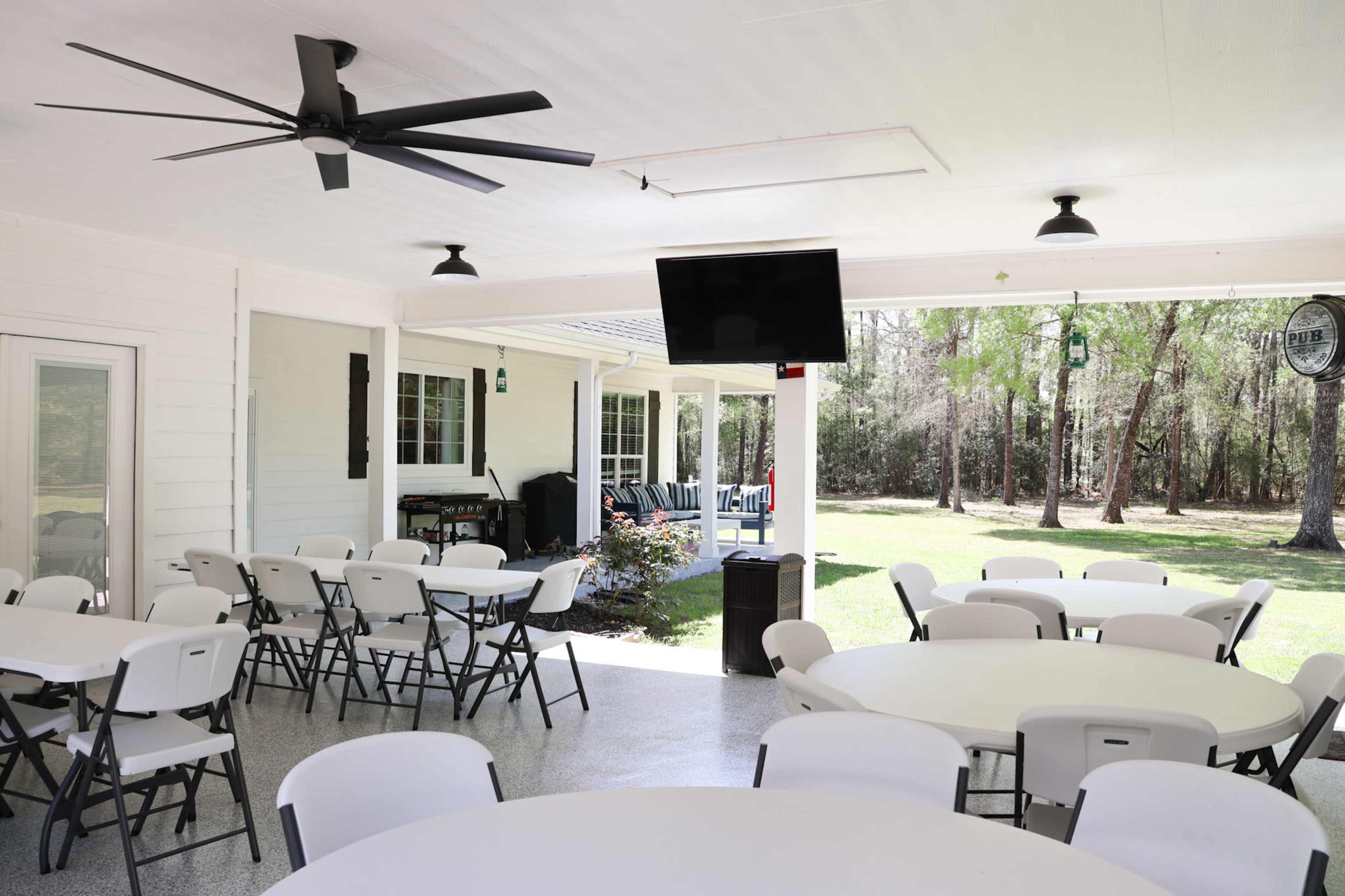 A spacious outdoor covered area with several circular tables and chairs, a ceiling fan, and a television mounted on the wall, overlooking a grassy yard with trees.